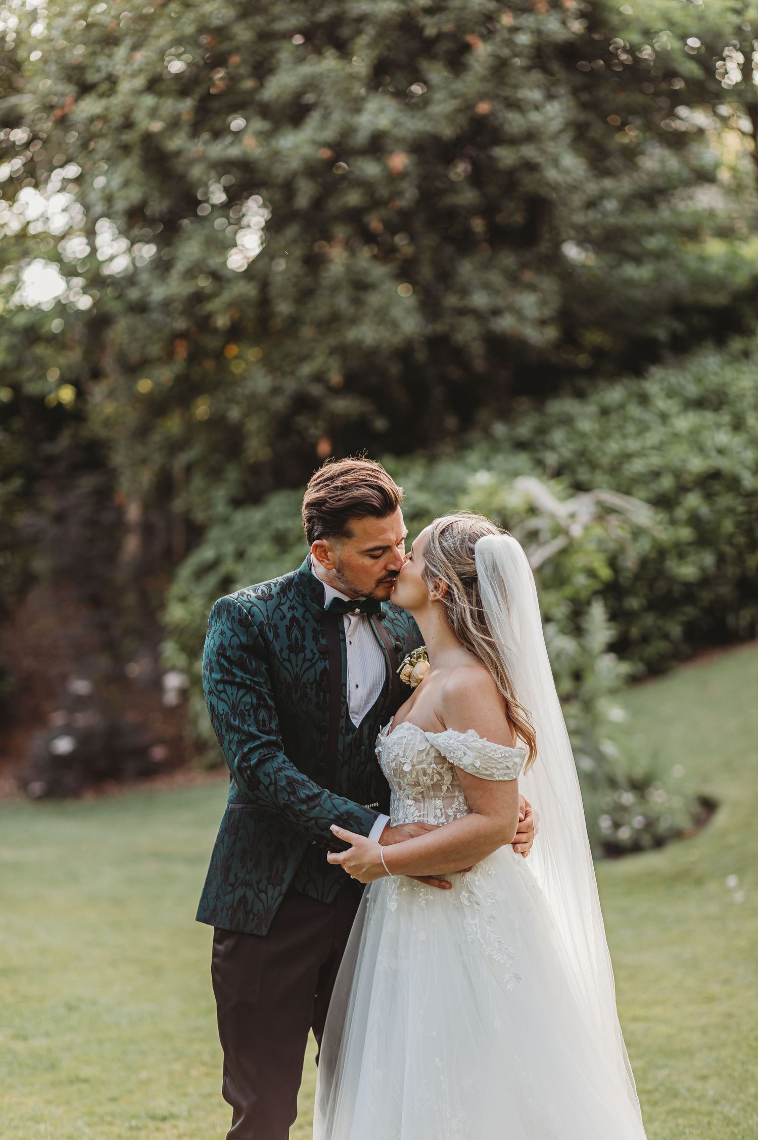 A romantic wedding couple embraces in an outdoor garden setting with stone walls and greenery.