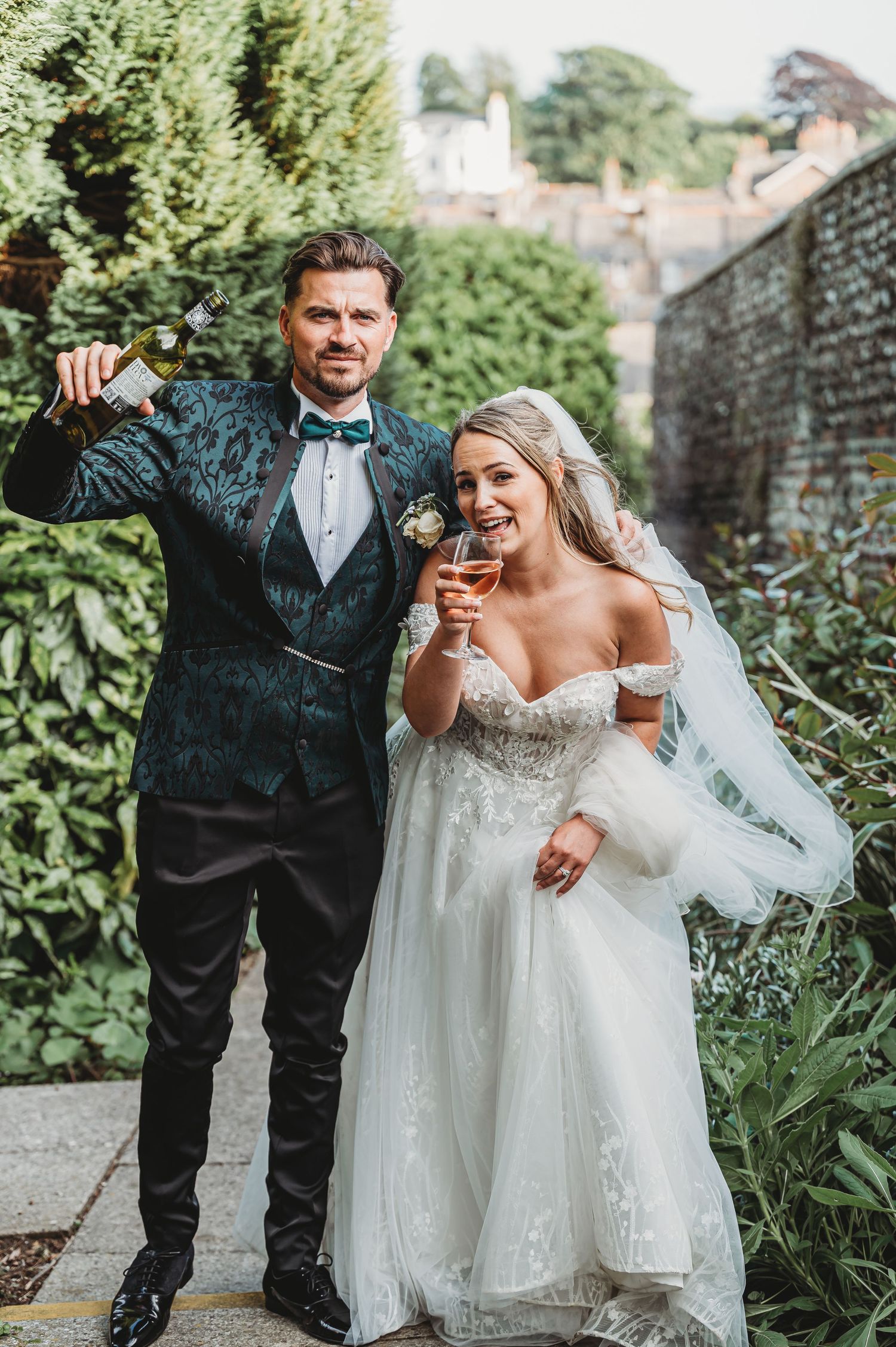 A joyful wedding couple shares a champagne toast in a garden setting surrounded by greenery.