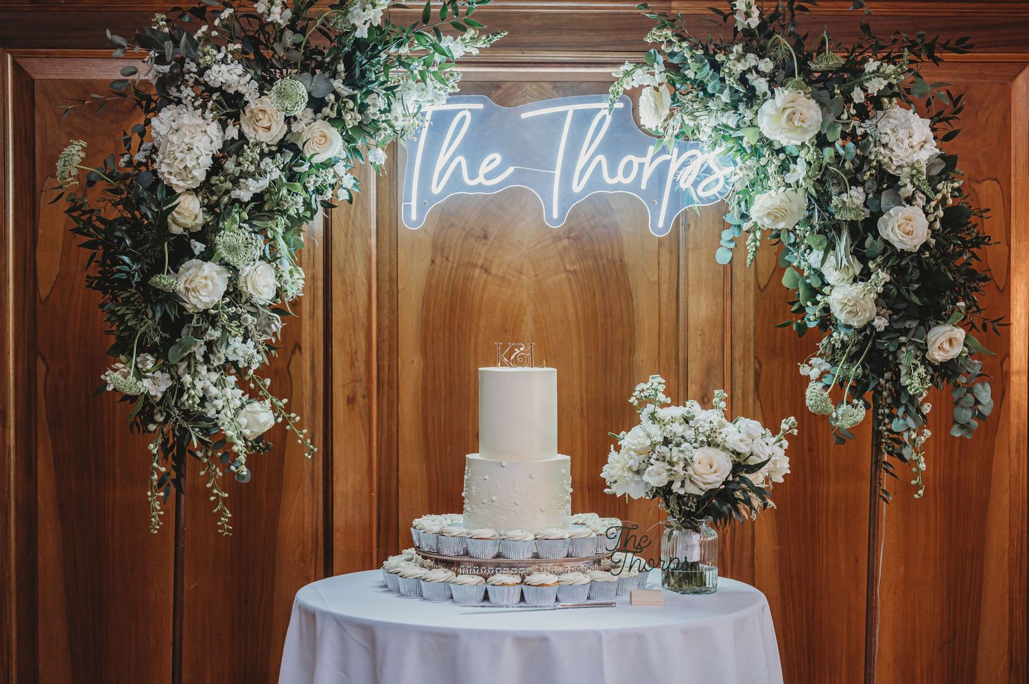 Wedding cake display with neon sign and white floral arch decorations.