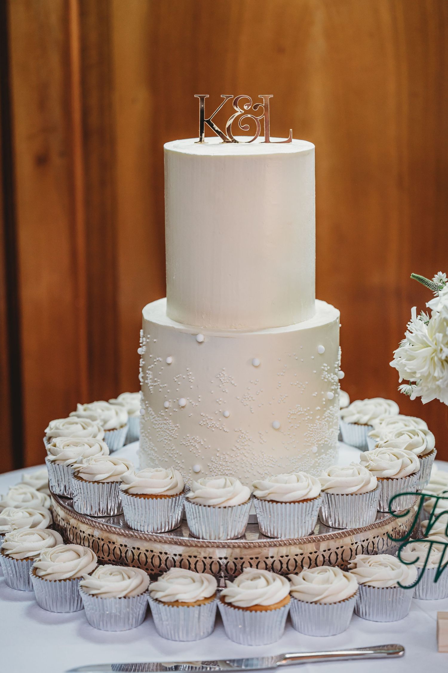 Elegant white two-tier wedding cake with cupcakes displayed on wooden stand.