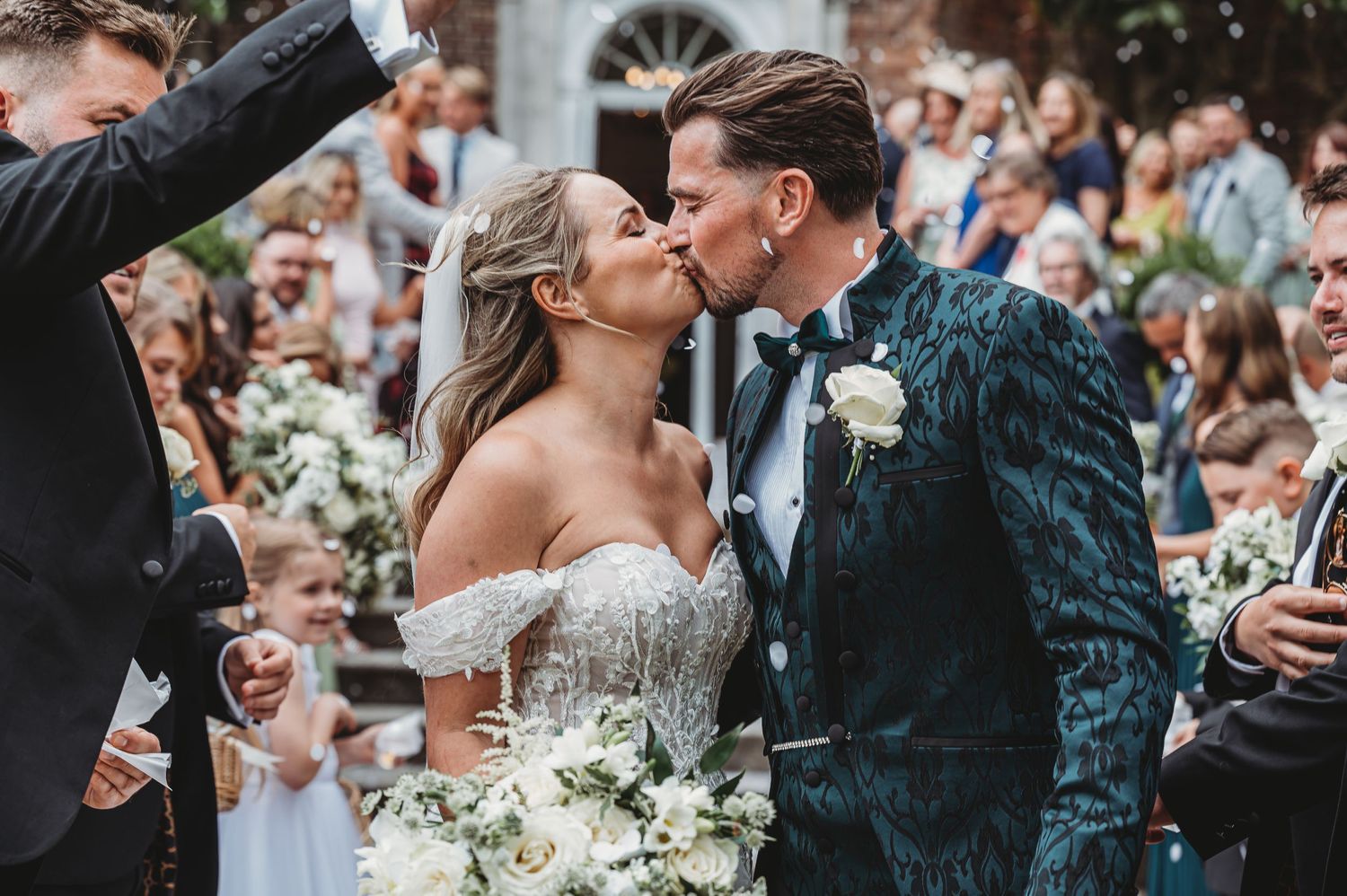 Romantic kiss surrounded by celebrating wedding guests and falling confetti.