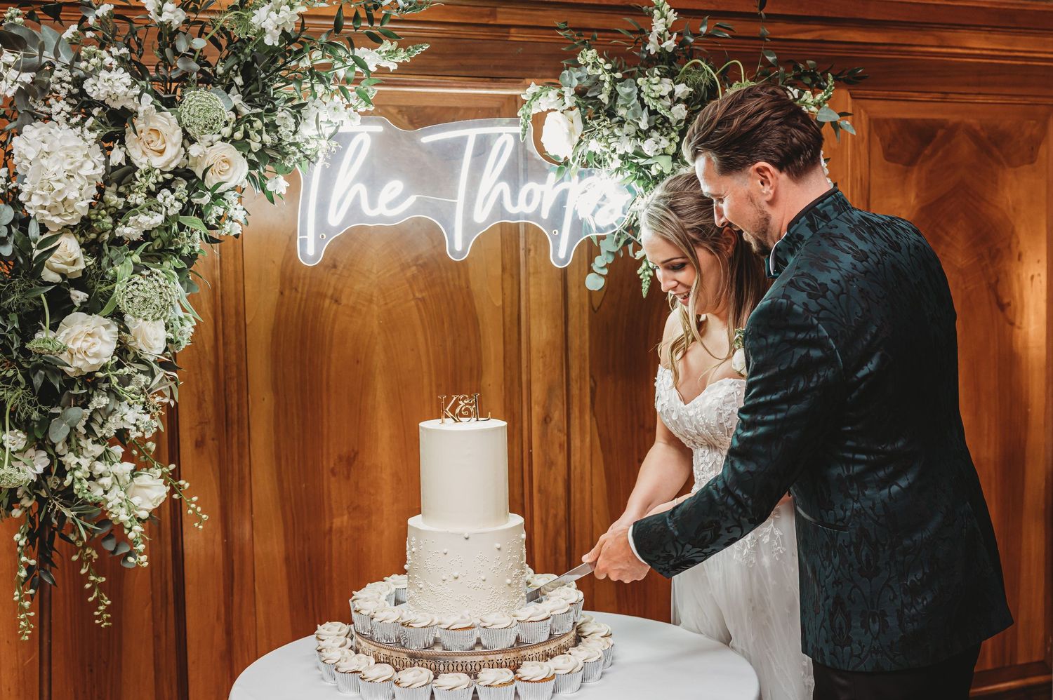 Wedding couple cuts their white tiered cake under a neon sign and floral arch.