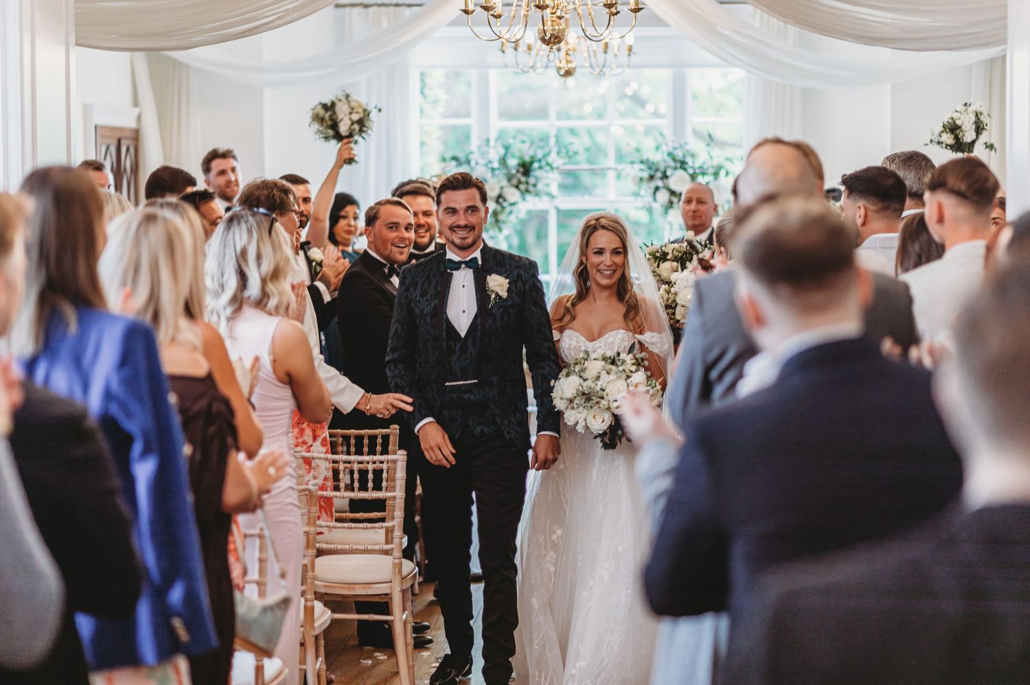 Wedding ceremony processional in an elegant white-decorated indoor venue with chandelier.