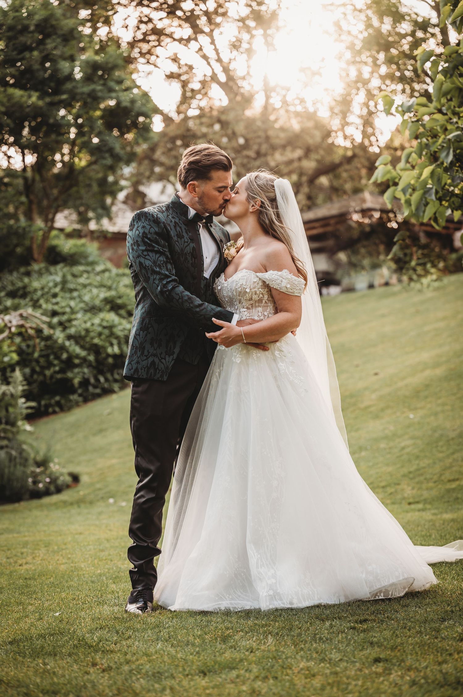 Newlyweds in elegant attire share a tender moment on manicured garden grounds at sunset.