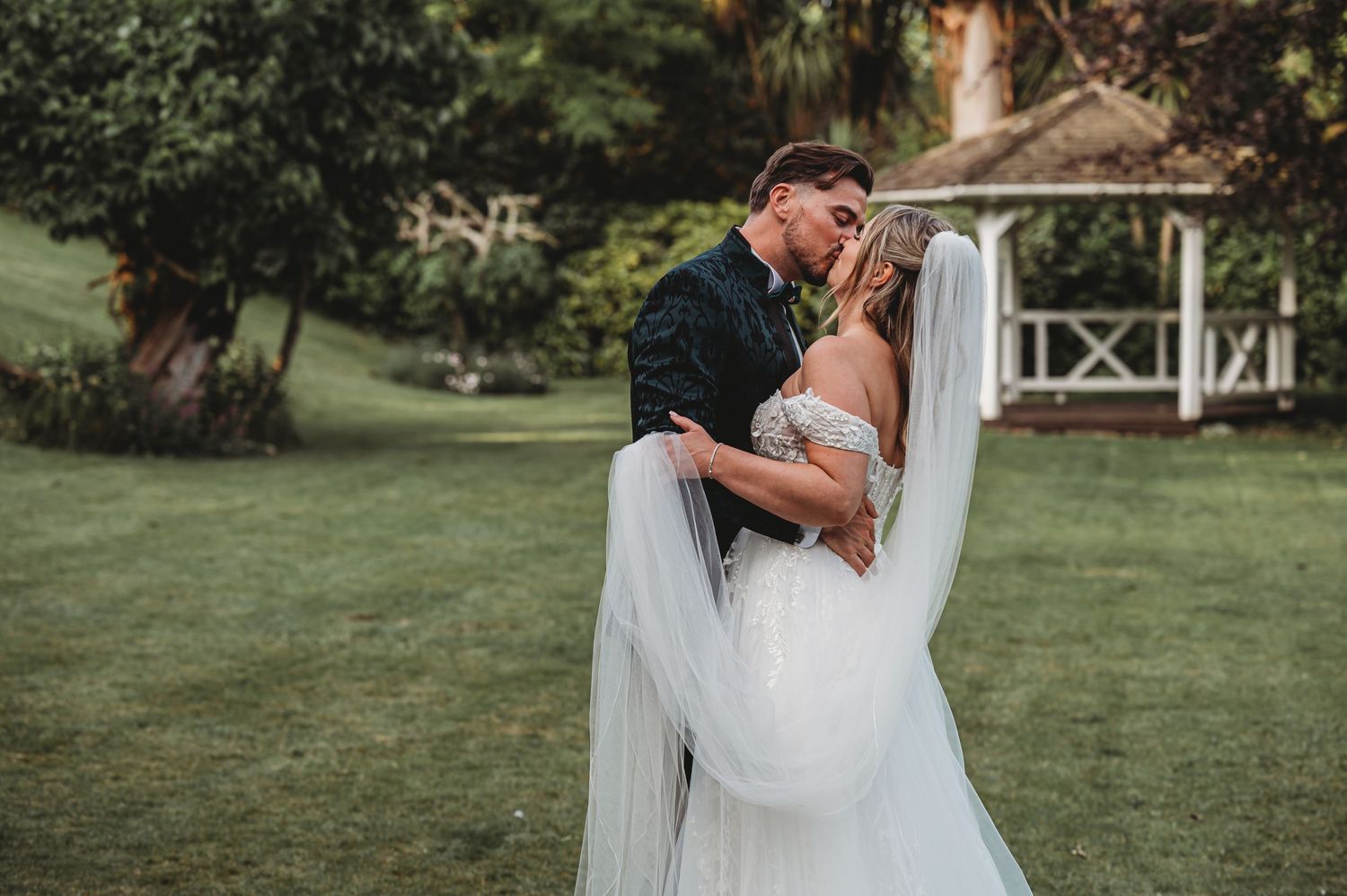 Romantic wedding couple embracing on a lush green lawn with a white gazebo in the background.