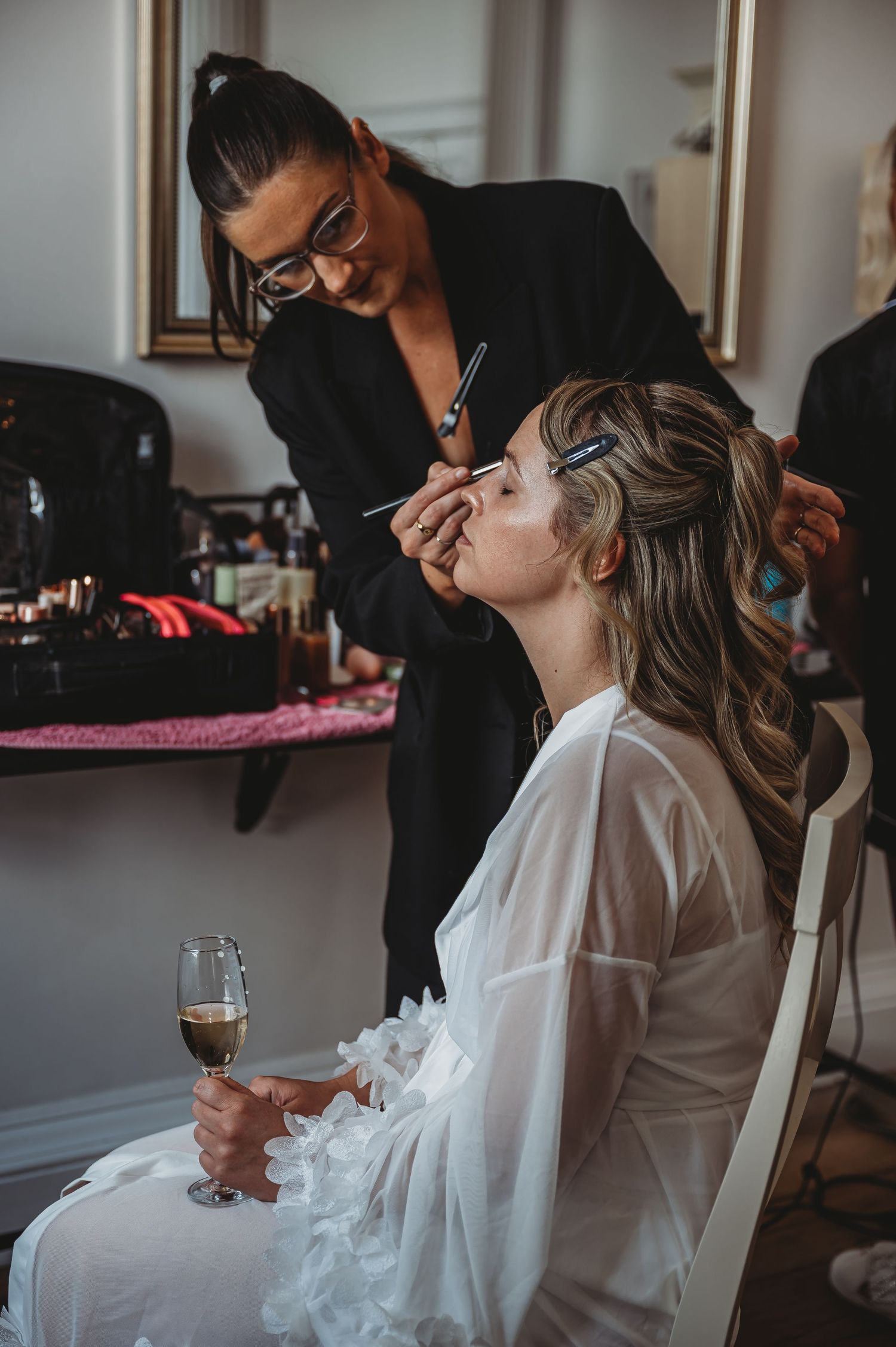 Makeup artist applying cosmetics to bride holding champagne during wedding preparations.