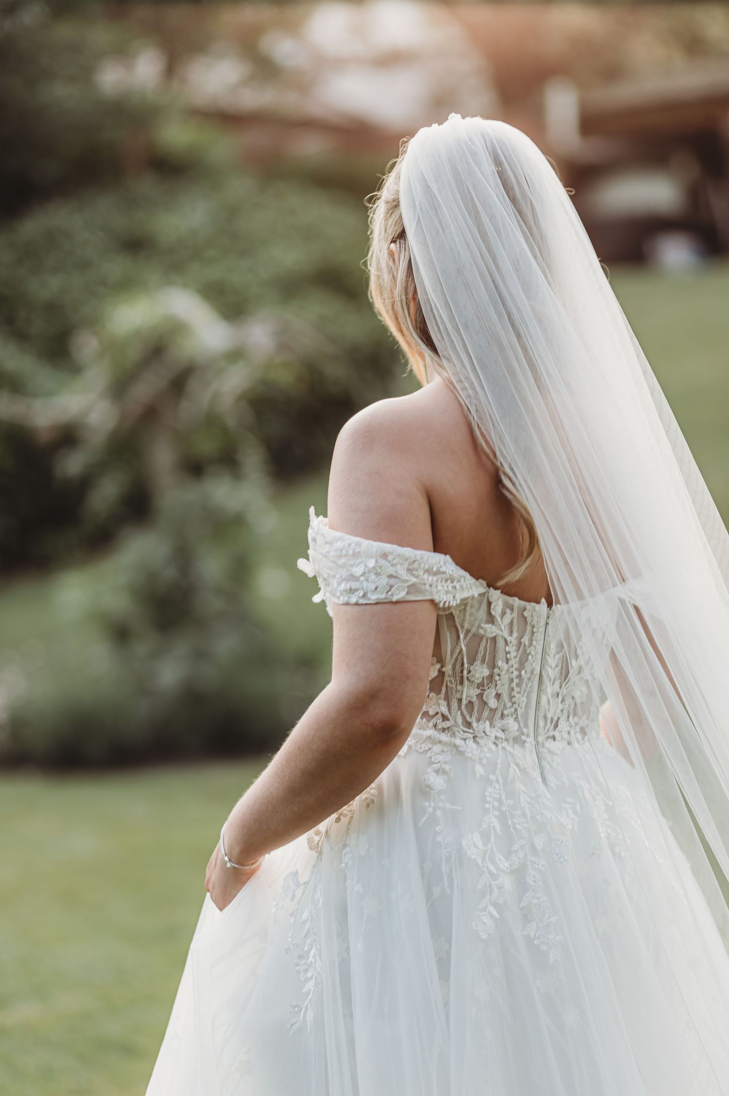 Back view of a wedding dress with off-shoulder lace details and flowing veil in an outdoor garden.