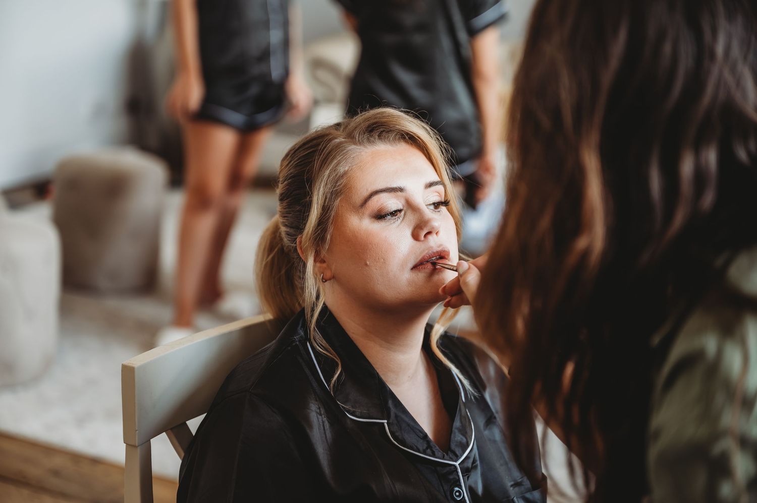 Makeup artist preparing bride for wedding day in stylish black satin robe.