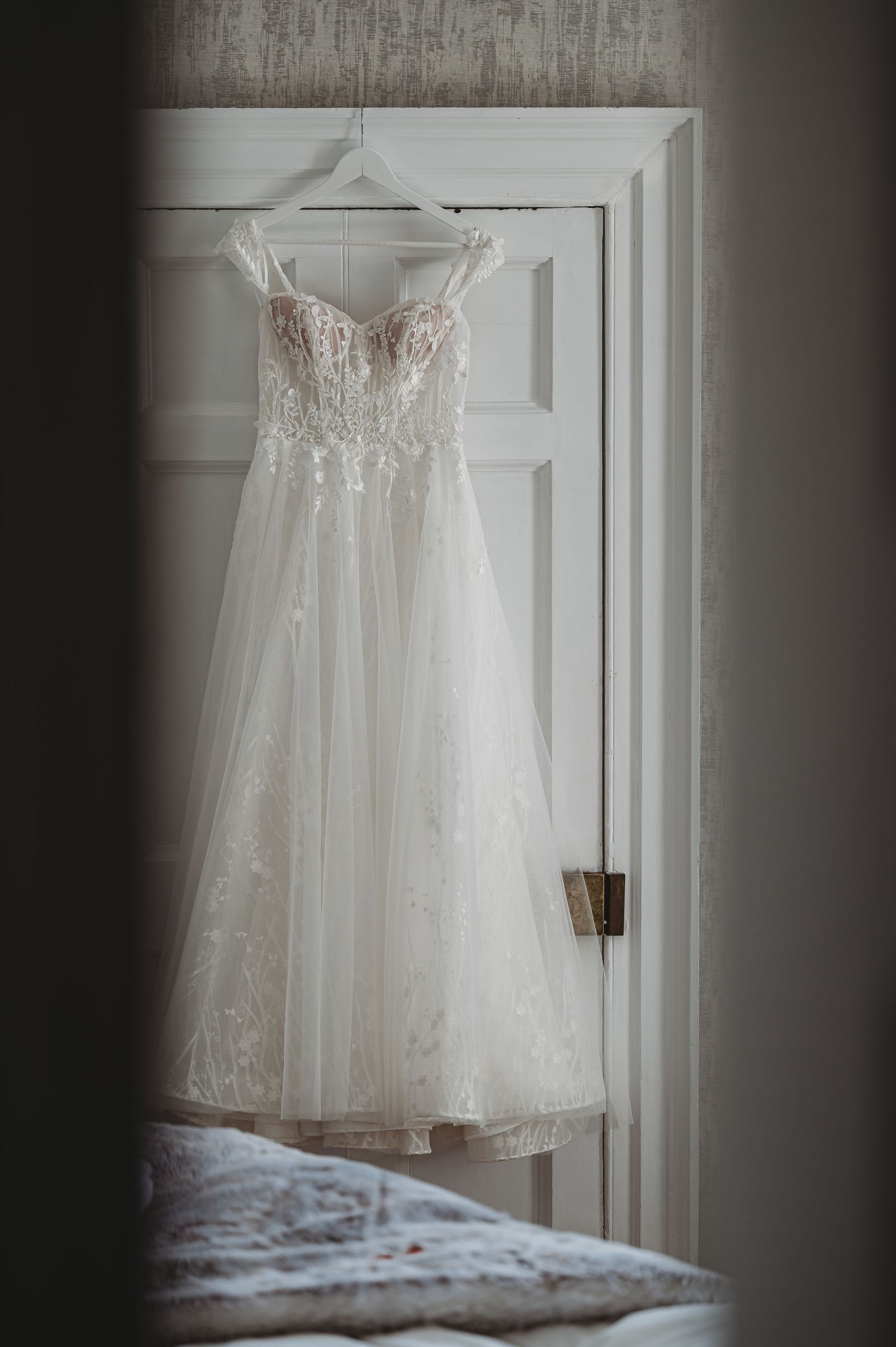 White lace wedding dress hanging on door in moody lighting.