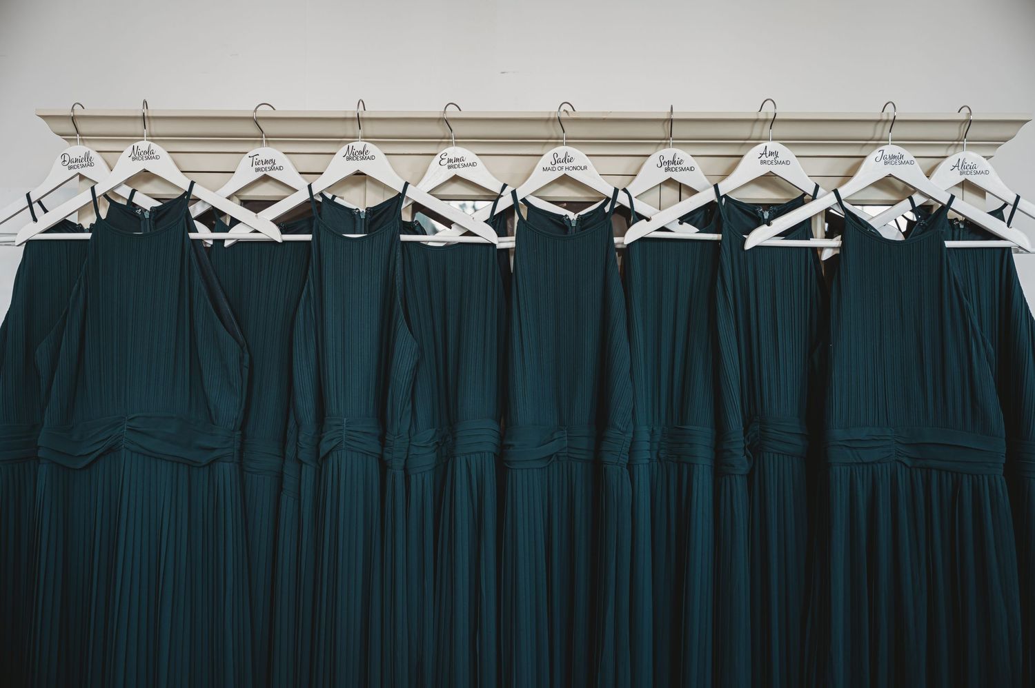 Row of emerald green bridesmaid dresses hanging on white hangers against light wall.