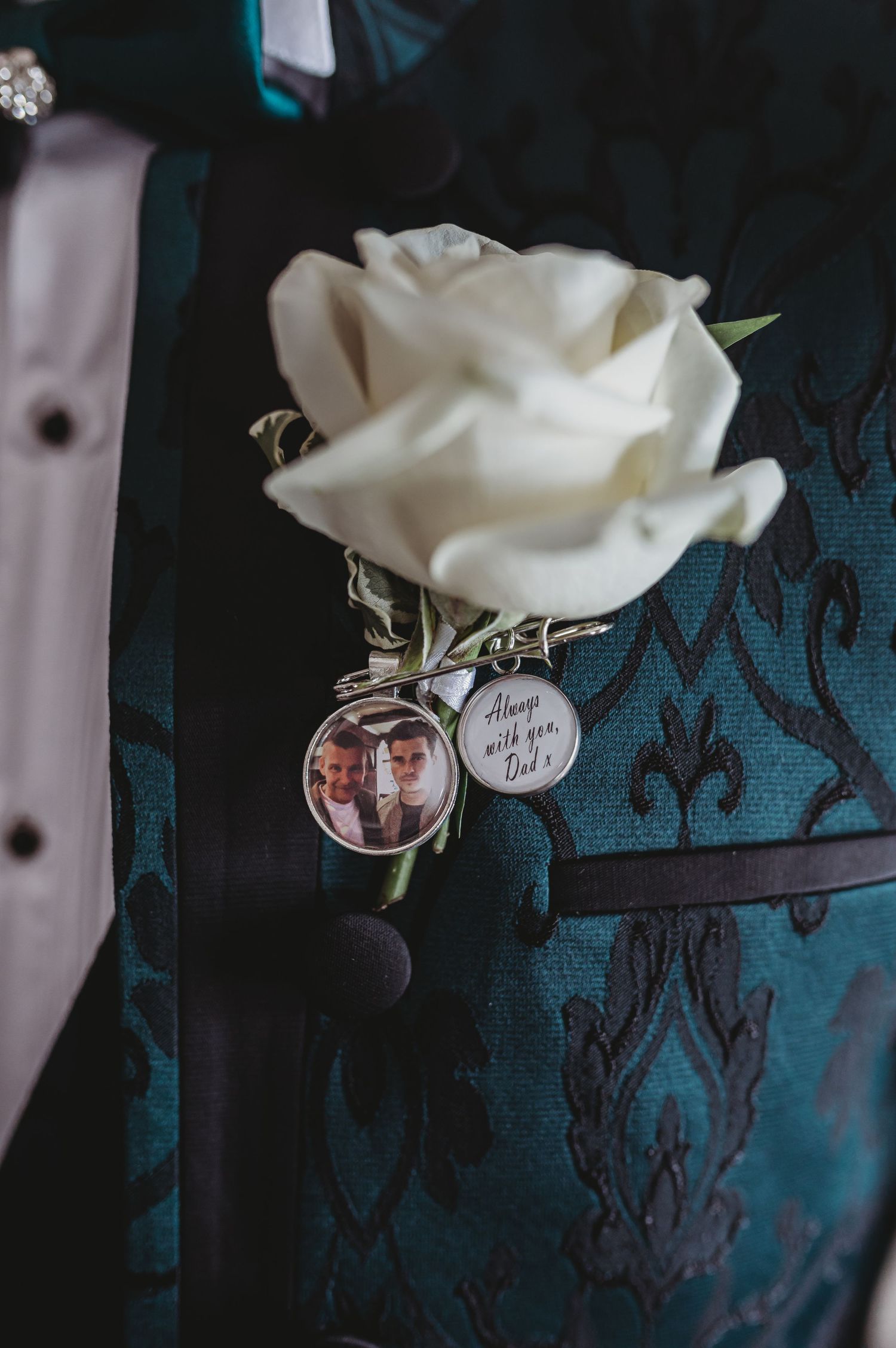 White rose boutonniere with decorative charm pin against dark patterned fabric.