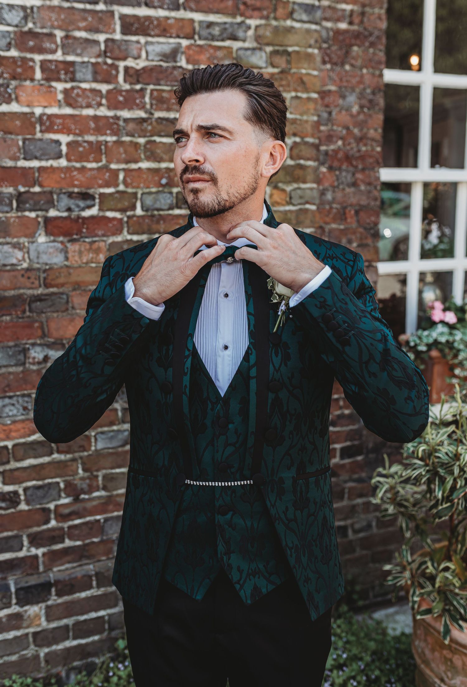 A groom adjusts his bowtie while wearing an elegant dark green velvet tuxedo against a brick wall backdrop.