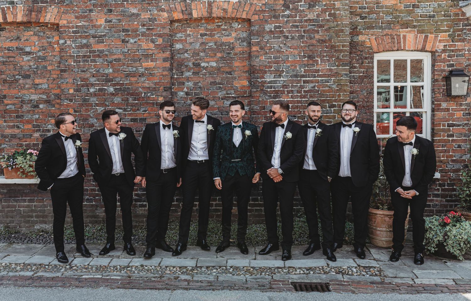 A wedding party of groomsmen in black tuxedos pose together against a rustic brick wall.