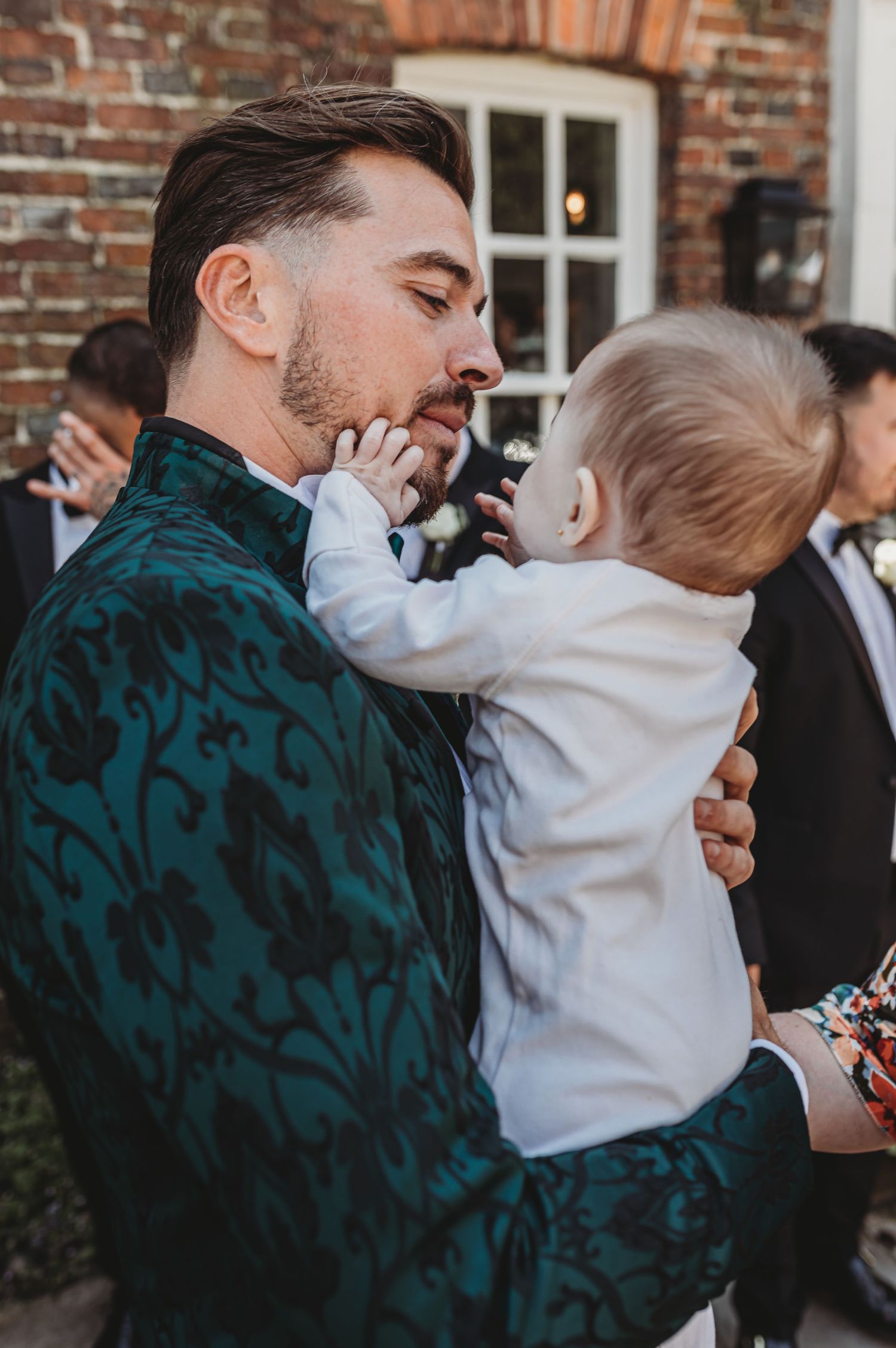 Sweet moment between adult in patterned jacket and baby in white outfit at formal event.