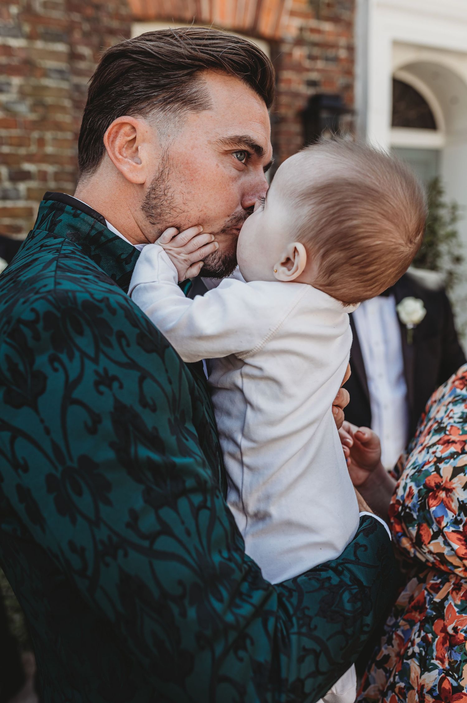 Person in emerald velvet jacket holding small baby in white outfit against brick wall.