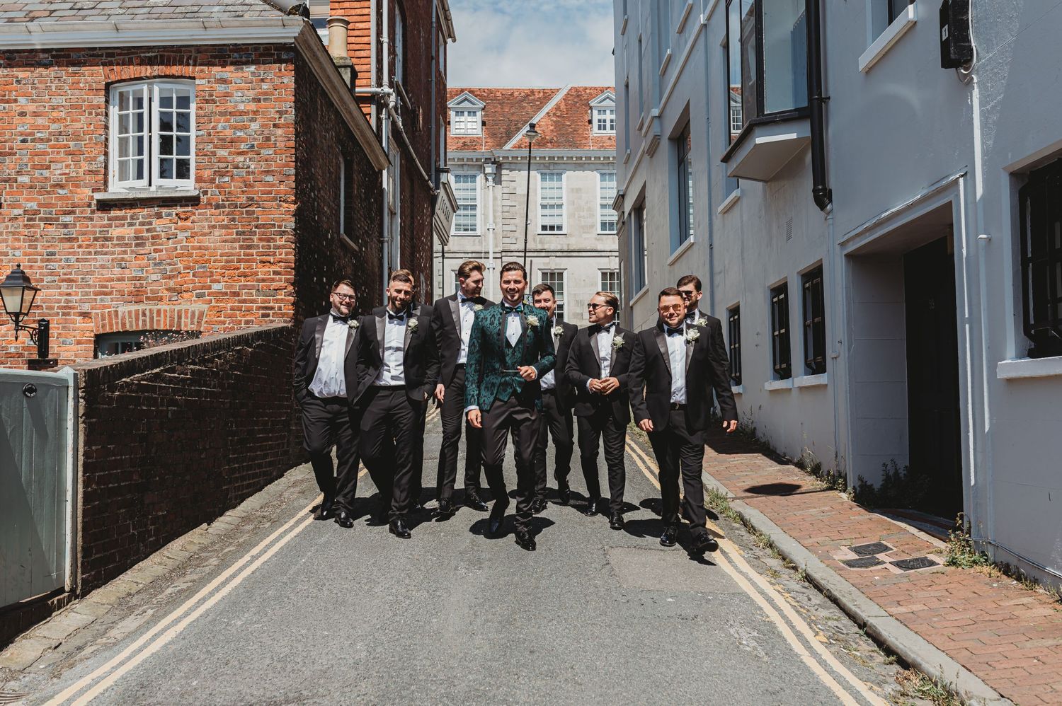 Groomsmen group photo on a narrow street between brick and blue buildings.