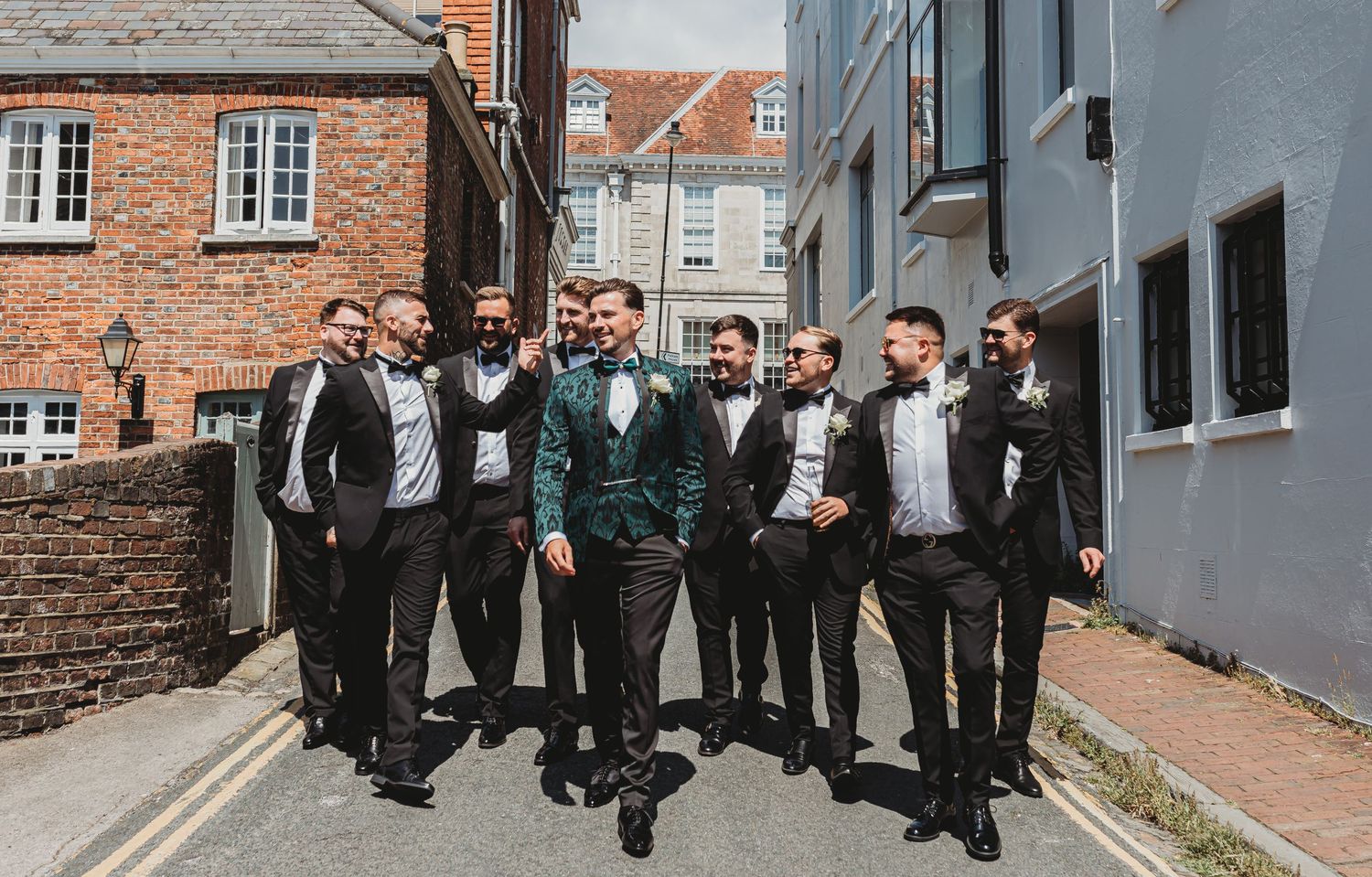 Wedding party posing outdoors in formal attire against brick building backdrop.