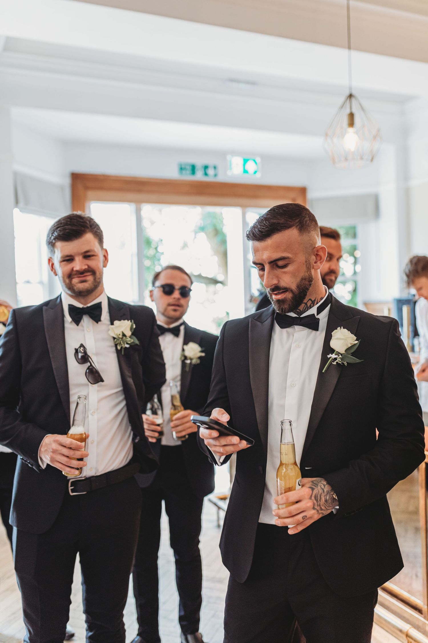 Two groomsmen in black bow ties holding drinks and chatting at wedding reception.