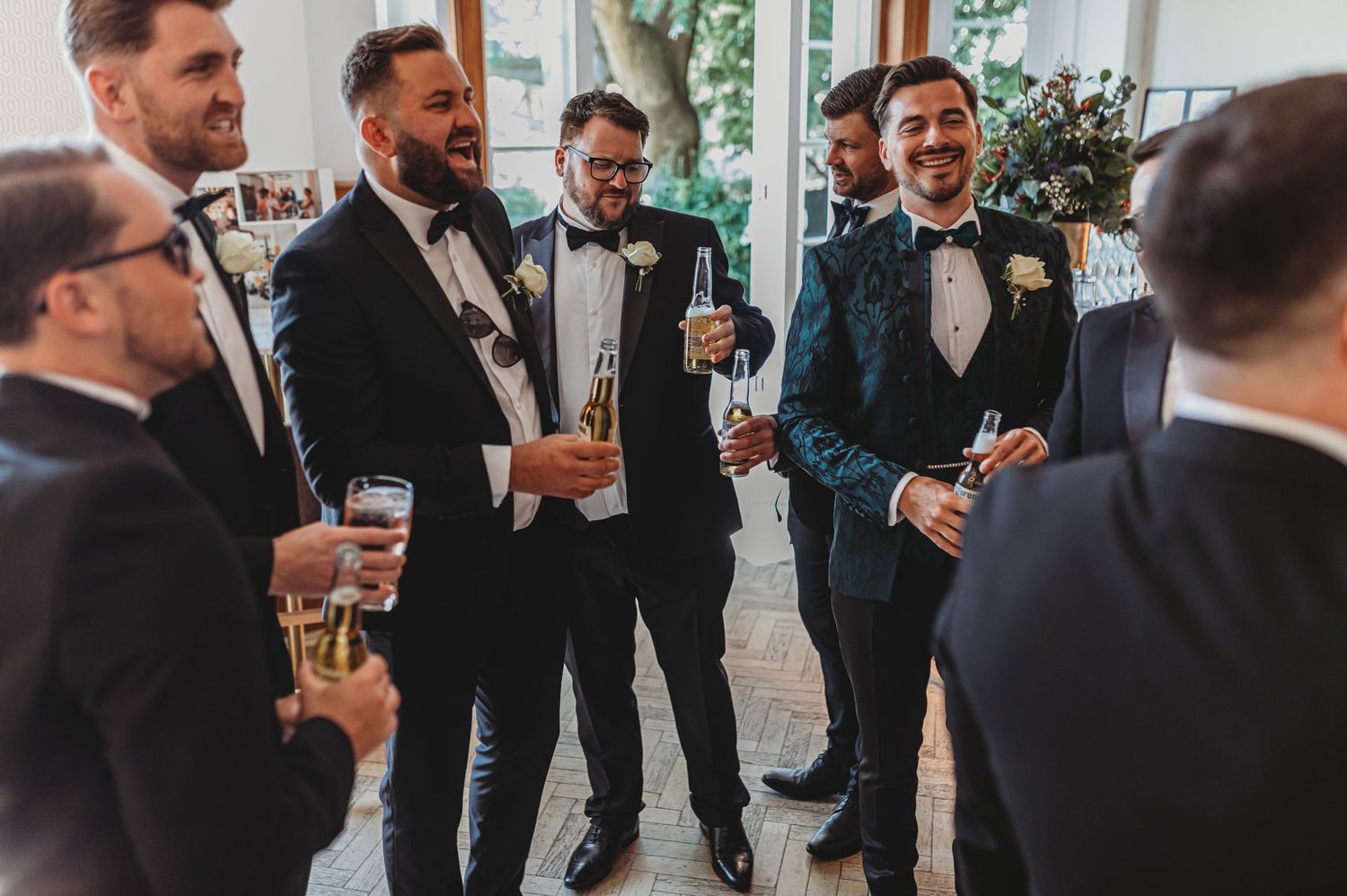 Wedding party of groomsmen laughing and celebrating with drinks in an elegant venue.