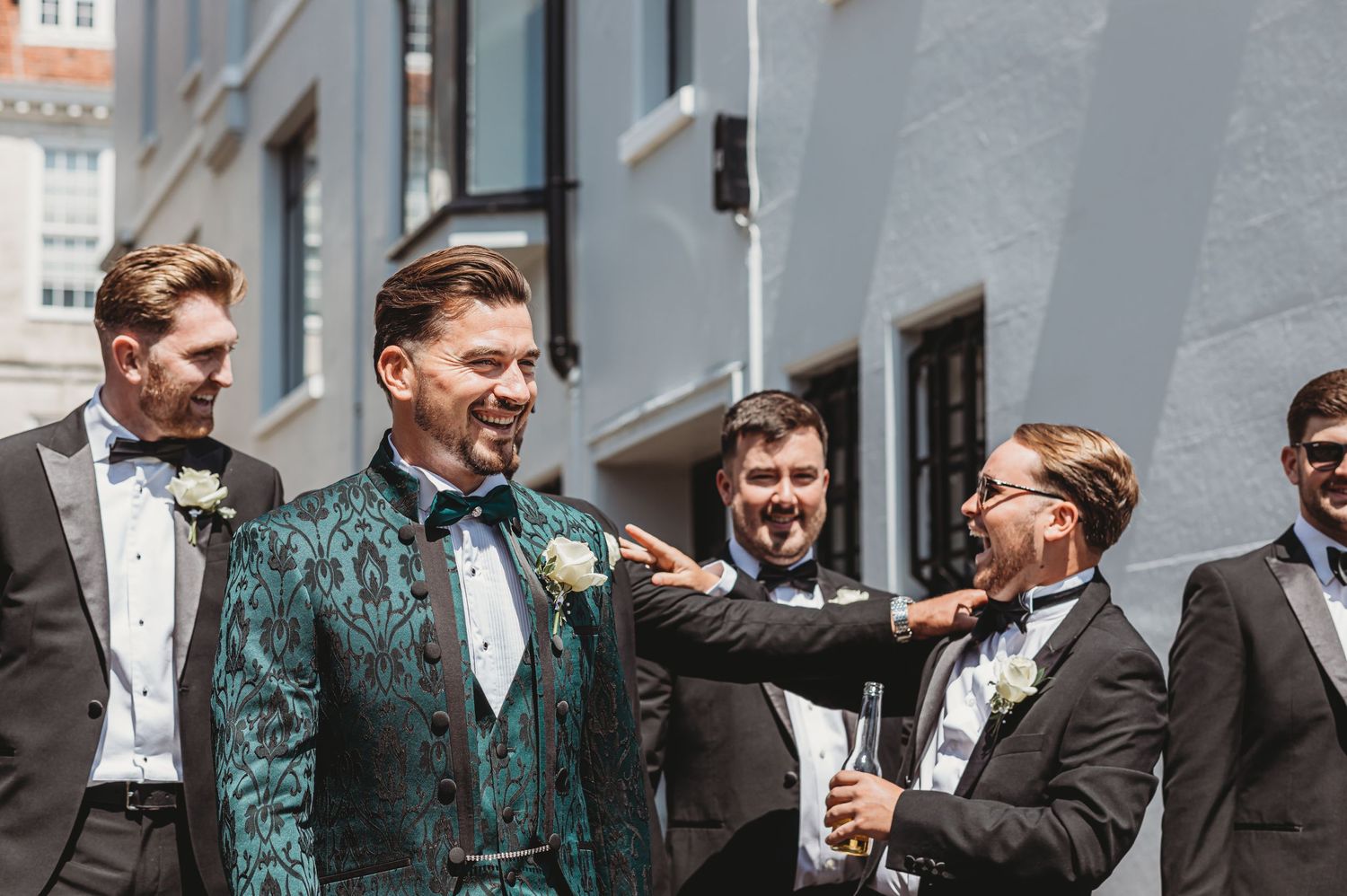 Wedding party in formal attire gathering outdoors with one member in a standout green velvet suit.