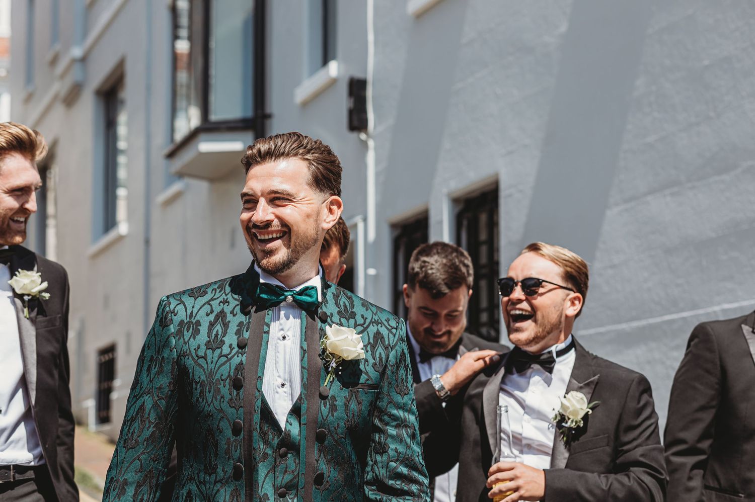 Groomsmen sharing a laugh outside while wearing formal attire including a striking emerald green jacket.