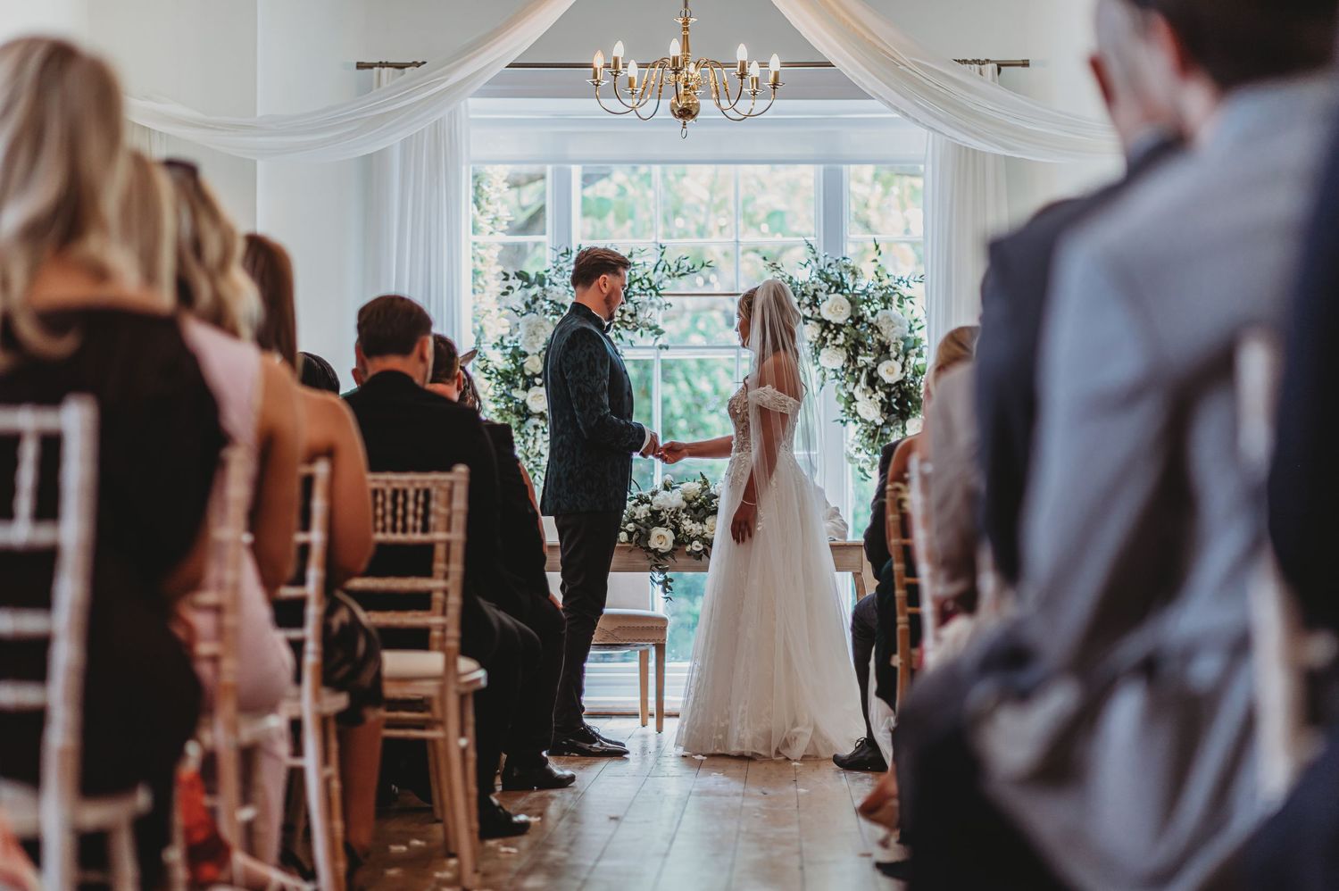 Wedding ceremony in bright indoor venue with white drapes and floral arch.