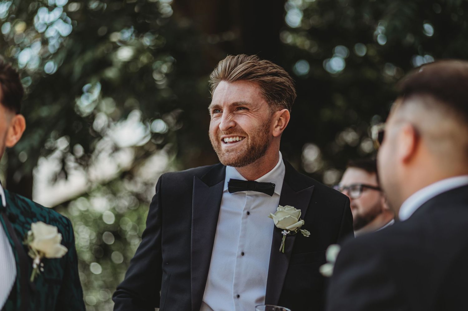 Groomsmen in formal attire sharing laughs during an outdoor wedding celebration.