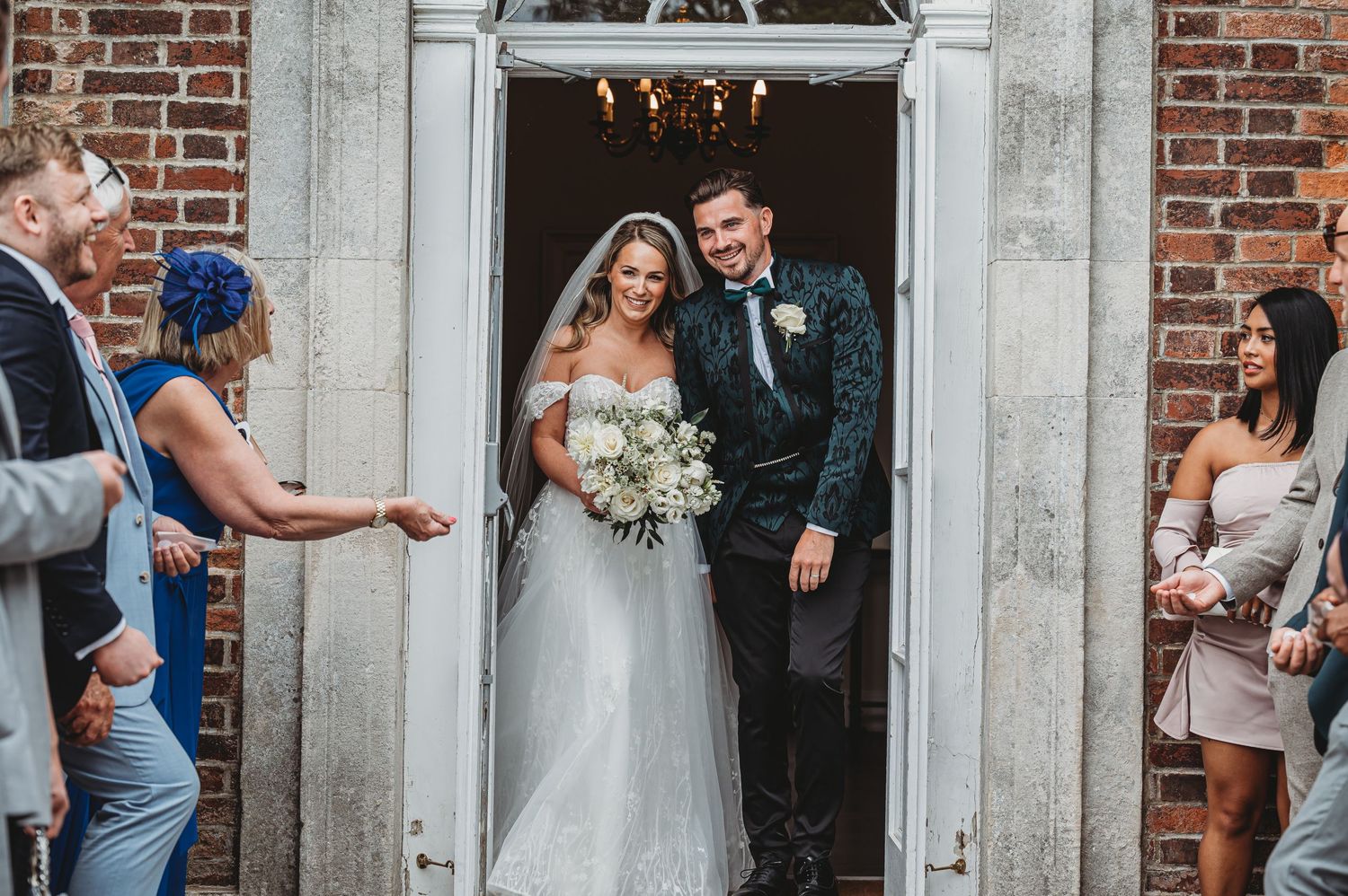 Wedding couple exits through ornate stone doorway while guests celebrate.
