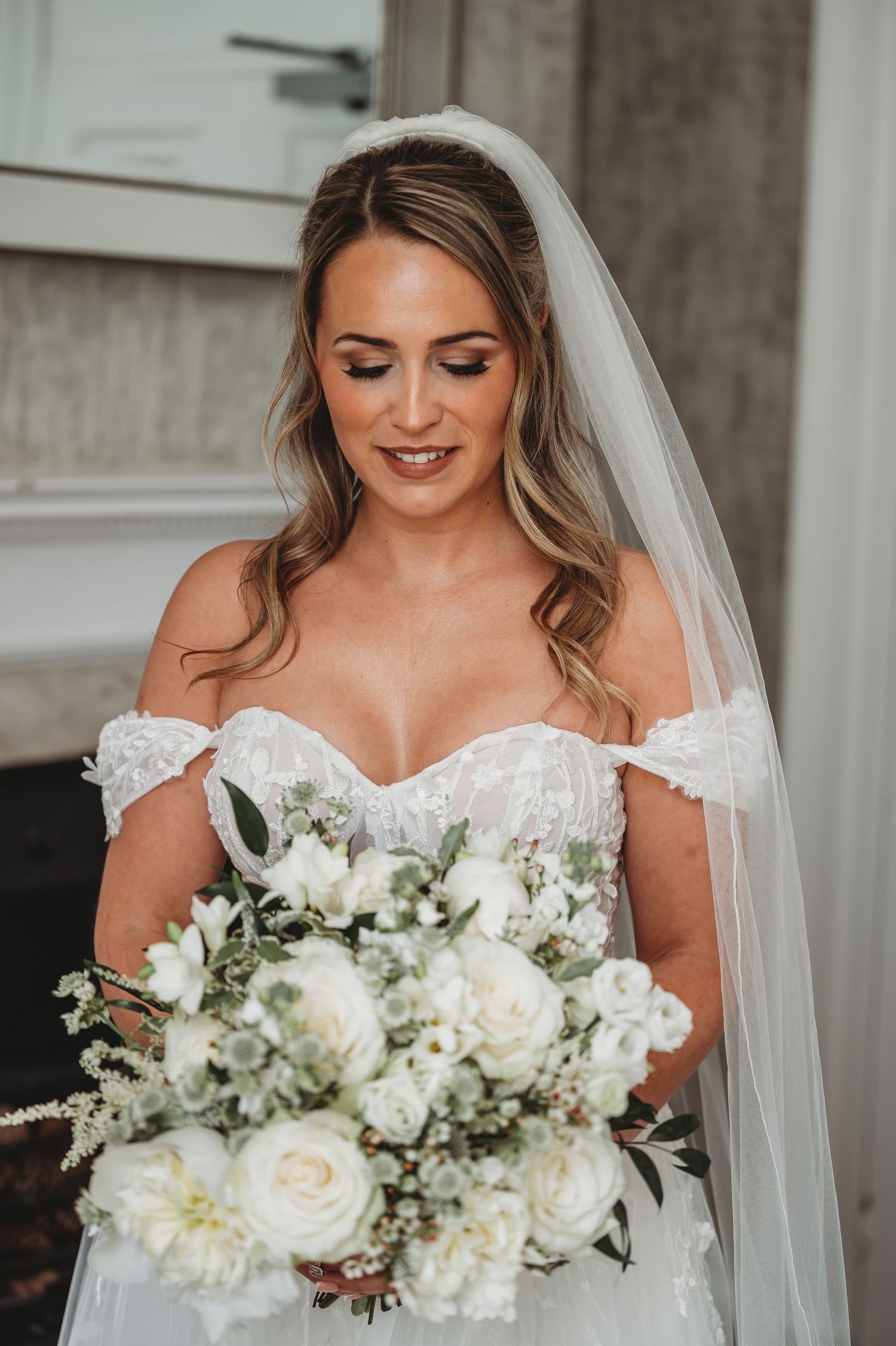 Bride holding white rose bouquet smiles while wearing off-shoulder lace wedding dress and veil.