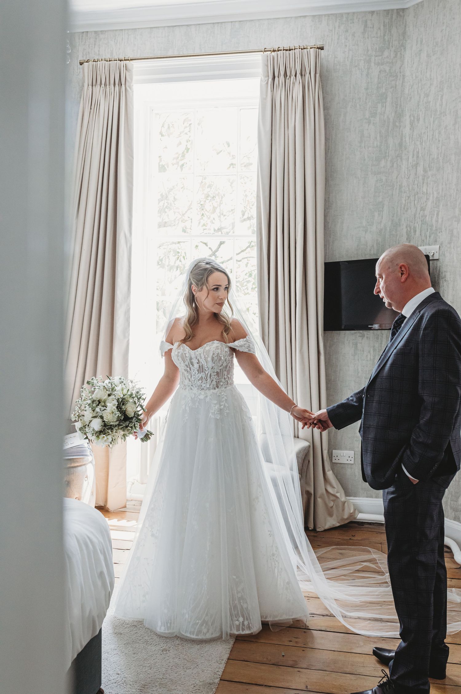Bride in lace wedding gown standing by window with suited man in elegant room.