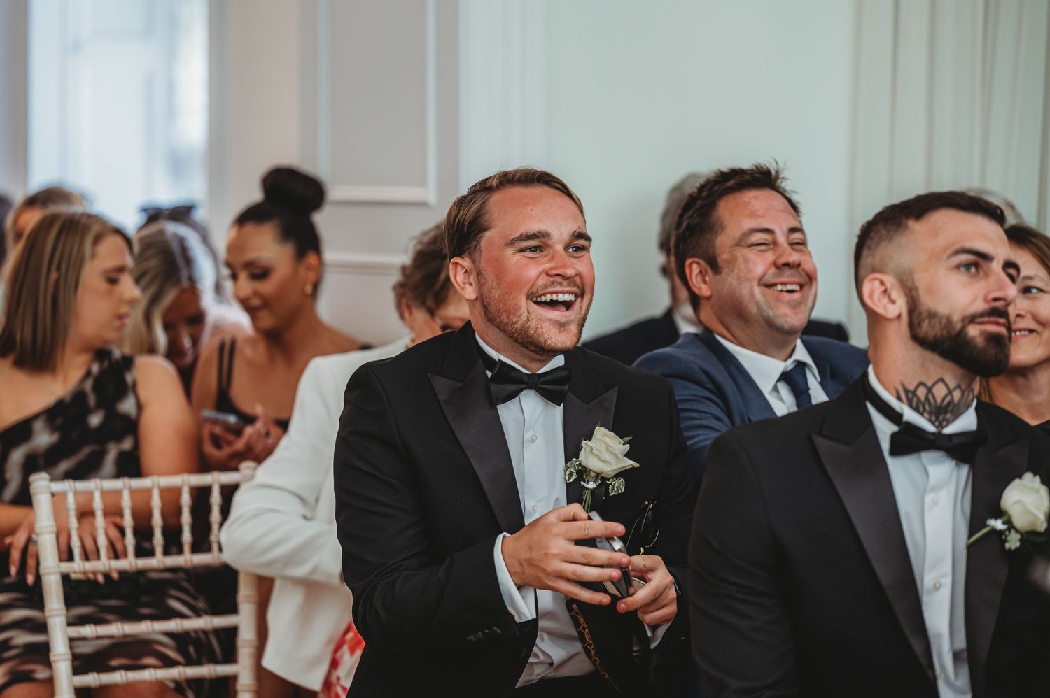 Wedding guests laugh and smile during a reception celebration while wearing formal attire in an elegant venue.