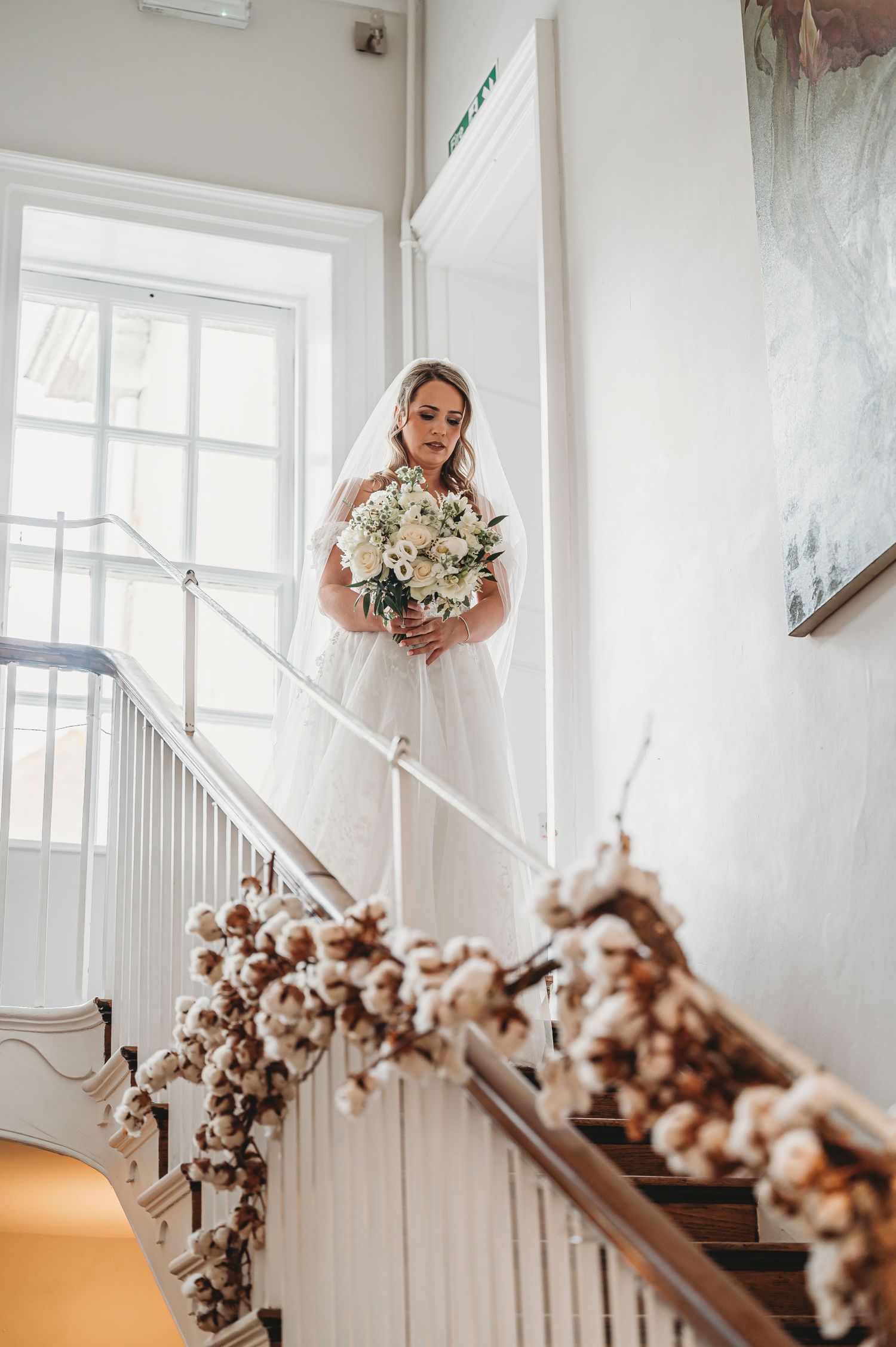 Bride in white gown on staircase with cotton branch garland and natural window light.