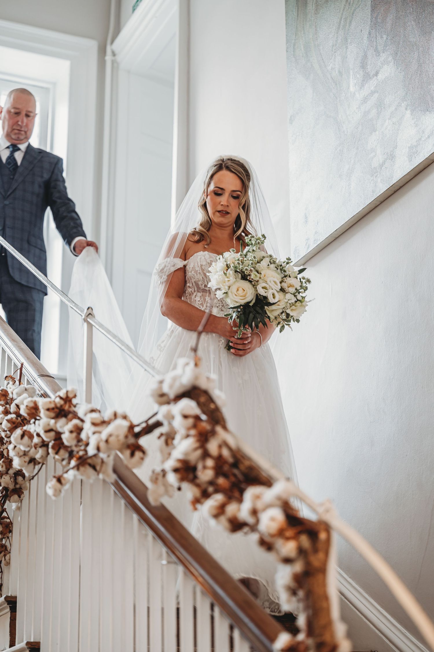 Bride in white gown holding bouquet on staircase decorated with cotton branch garland.