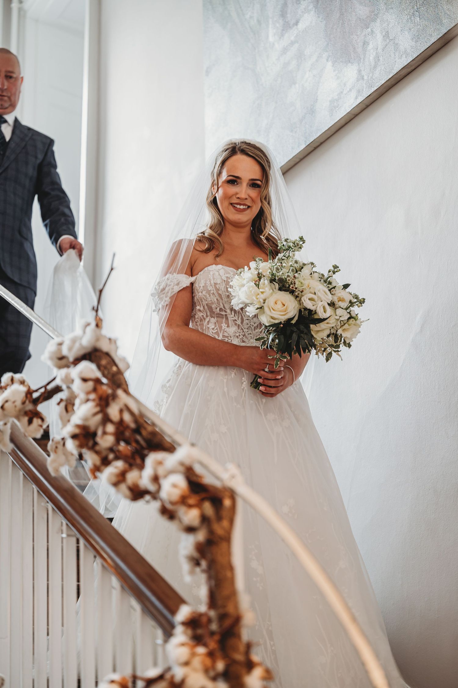 Elegant bride in white gown descending decorated staircase with cotton branch garland.