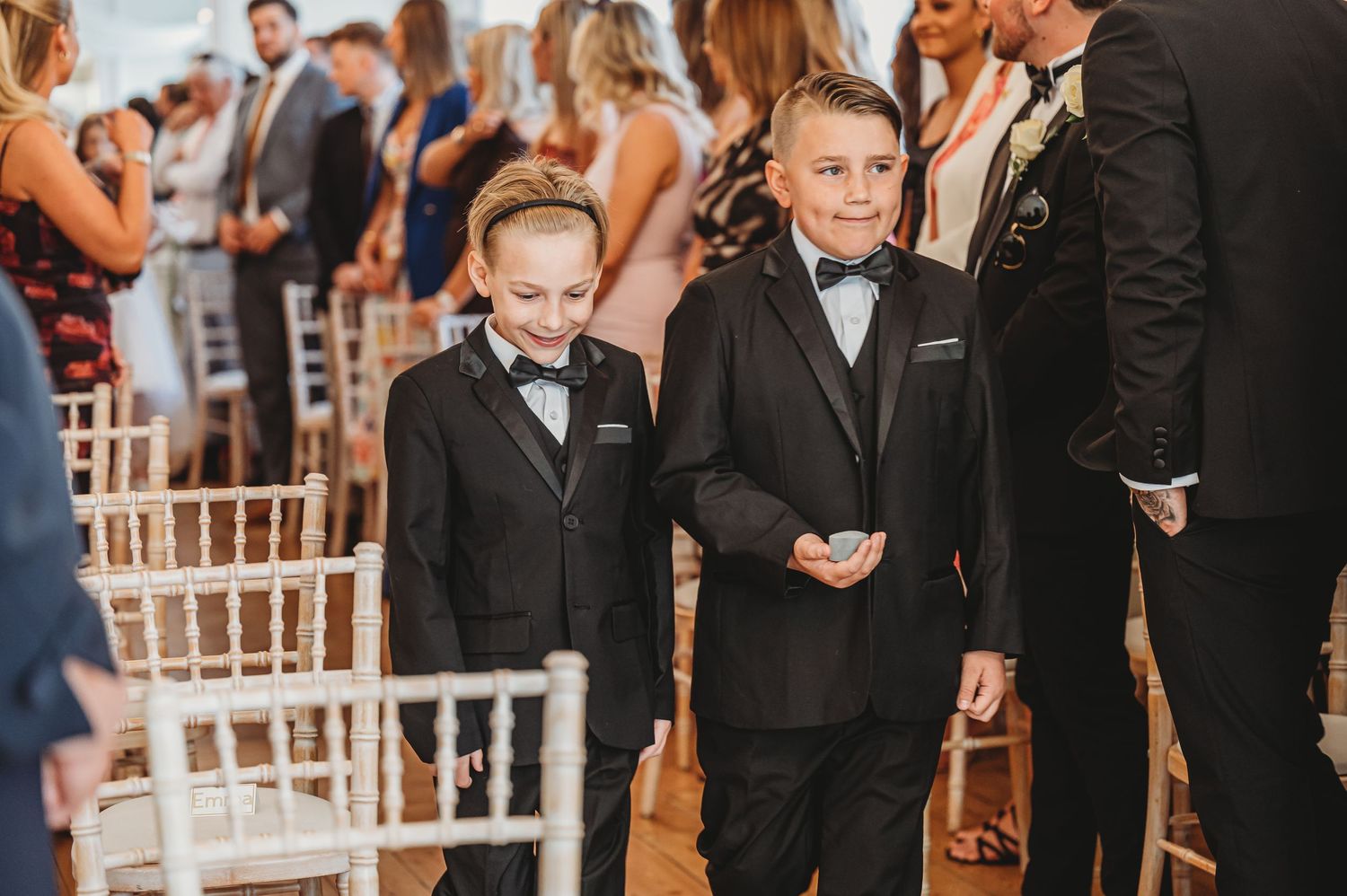 Young ring bearers in black tuxedos and bow ties standing during wedding ceremony.