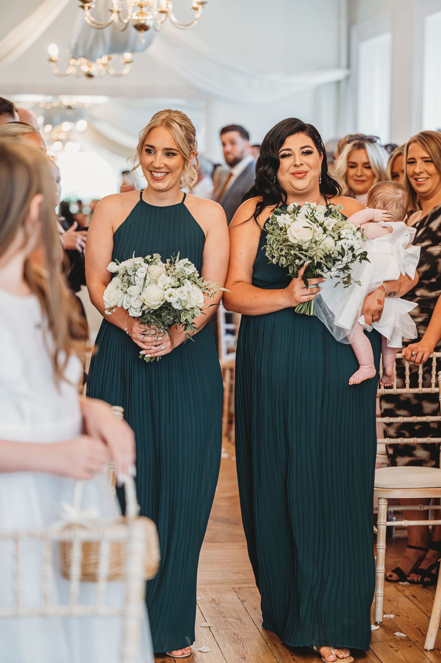 Bridesmaids in teal floor-length dresses holding white bouquets during wedding ceremony.
