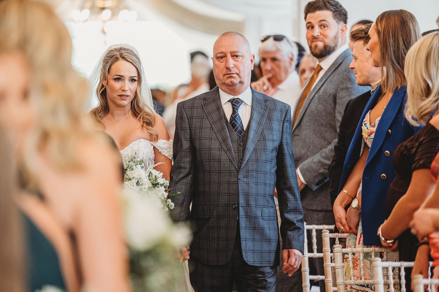 Indoor wedding ceremony with guests standing in formal attire and elegant decor.