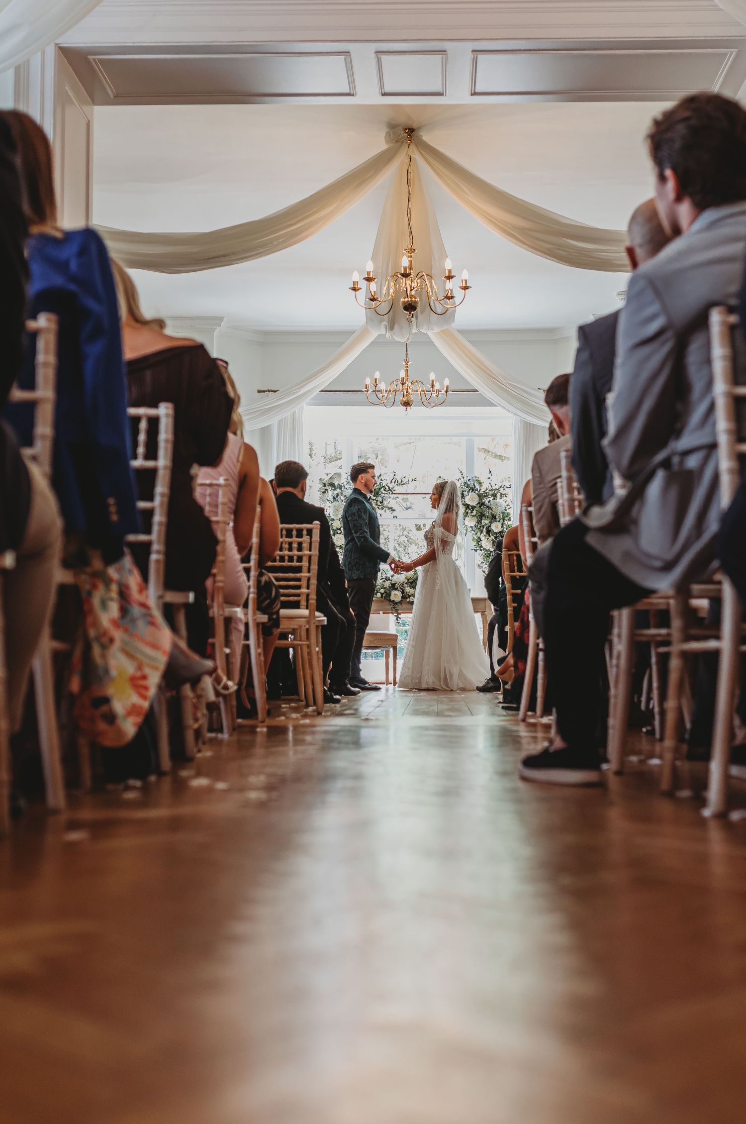 Wedding ceremony view down the aisle with white draping and chandeliers in an elegant indoor venue.