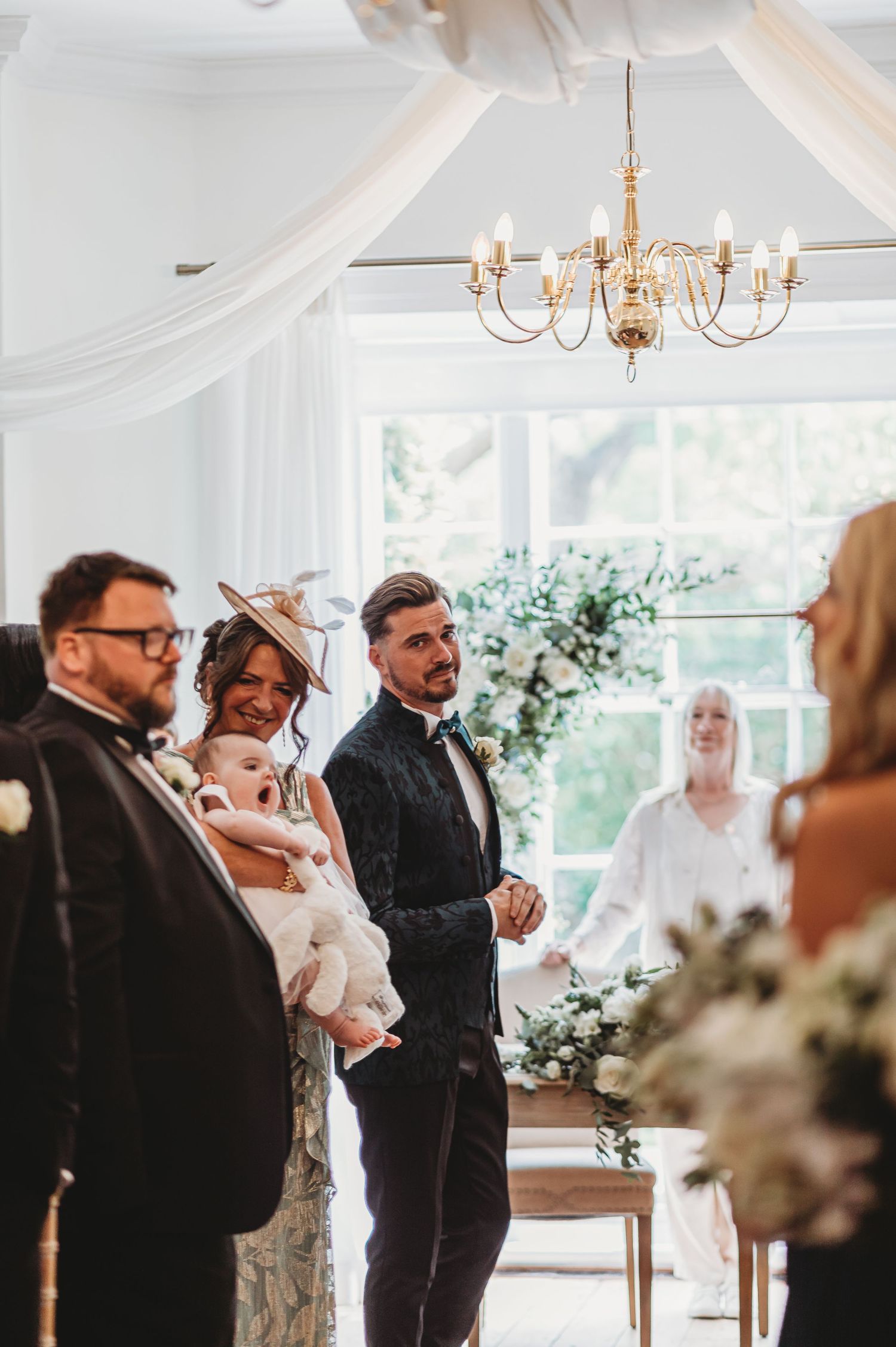 Wedding ceremony captured in an elegant white-draped venue with a crystal chandelier overhead and floral decorations.