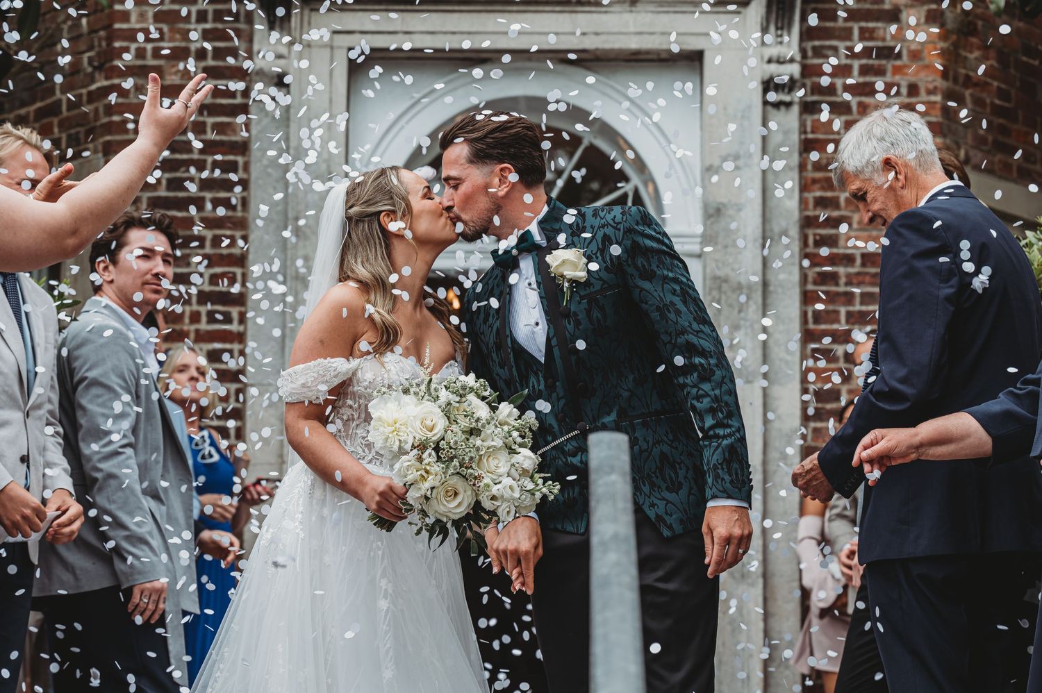 Romantic wedding exit under historic brick archway with guests throwing confetti.