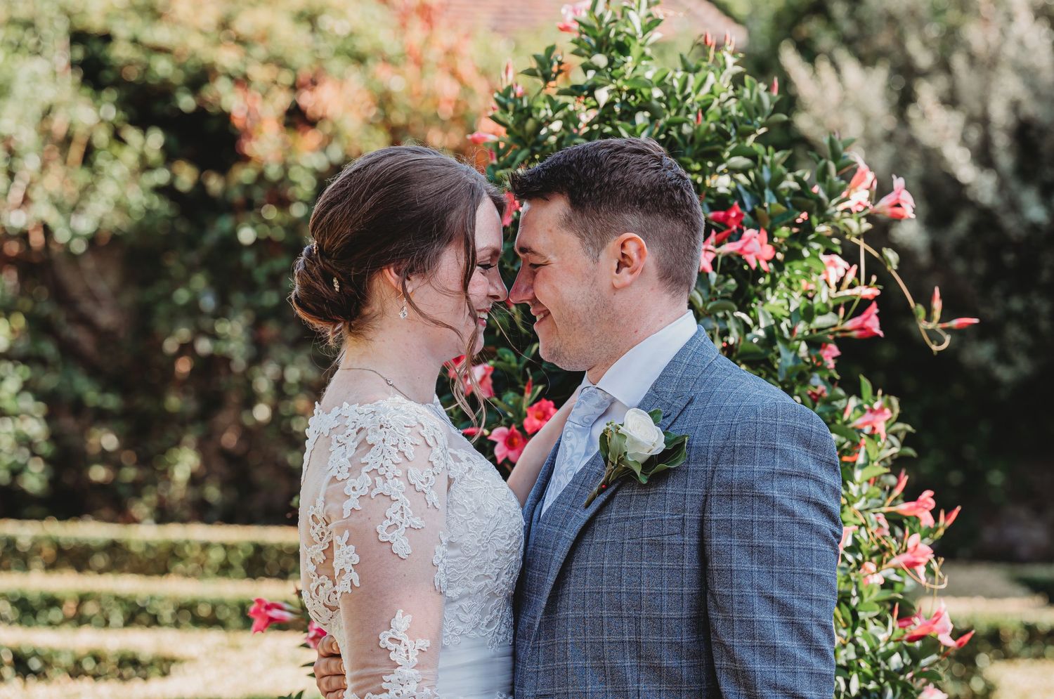 Couple in wedding attire share an intimate moment in a garden setting with pink flowers and greenery.