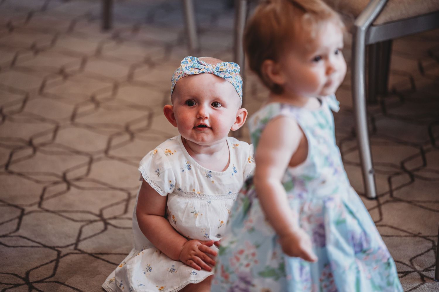 Two young children in summer dresses stand on a patterned tile floor, one smiling while wearing a headband.