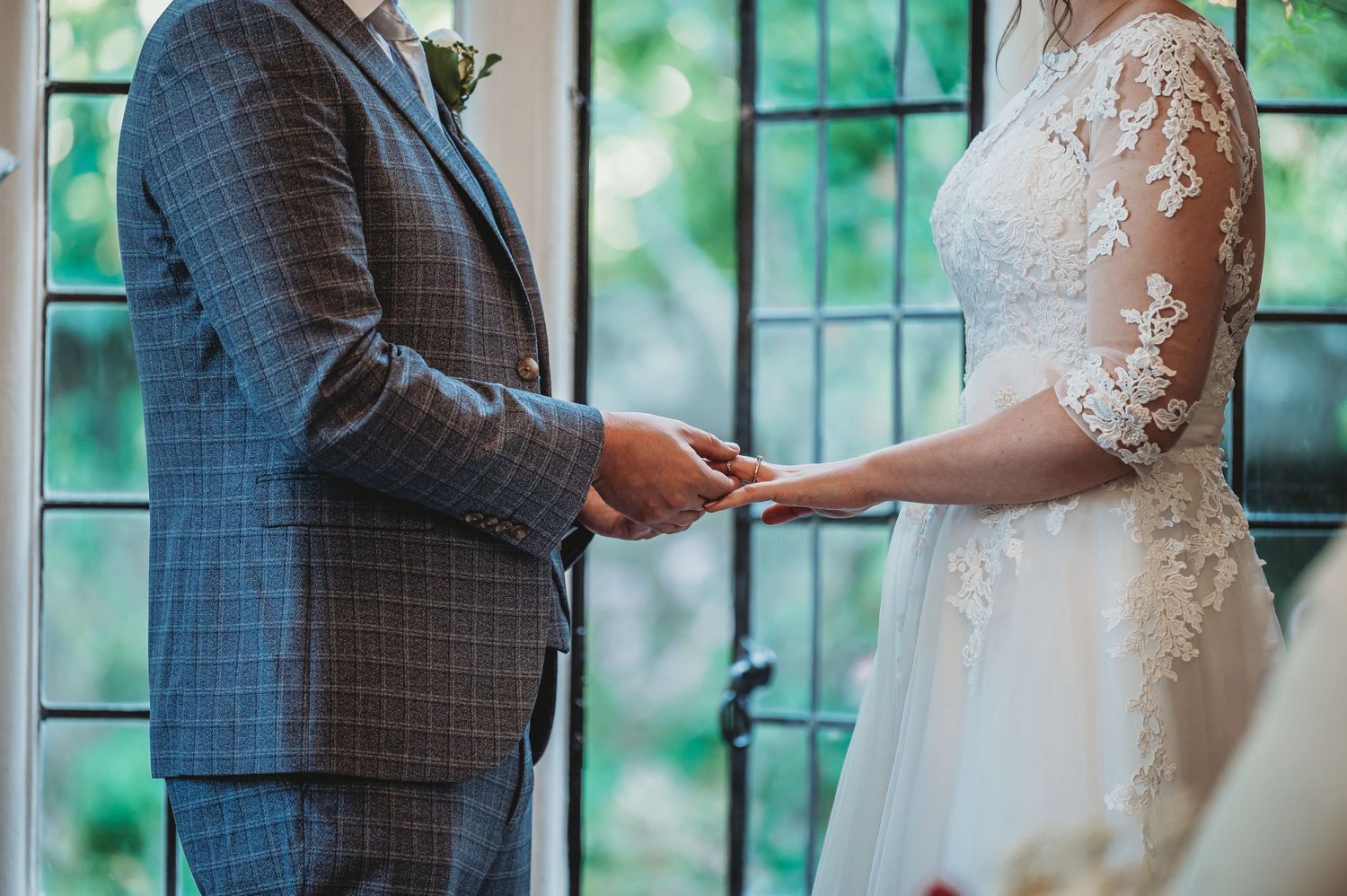 A couple exchanges rings during an intimate wedding ceremony by a window with green foliage in the background.