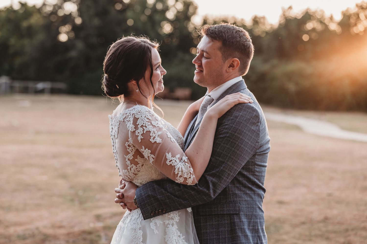 A loving couple embraces during golden hour on their wedding day, with the bride in lace and groom in grey suit.