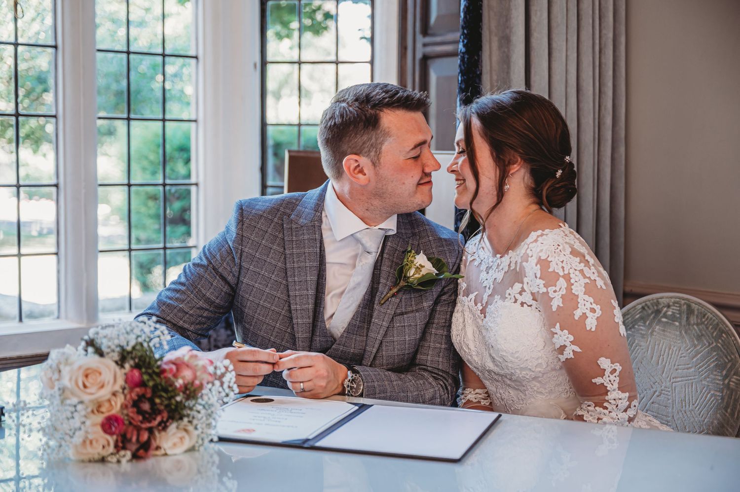 A couple in wedding attire shares an intimate moment while sitting at a table with autumn flowers and signing documents.