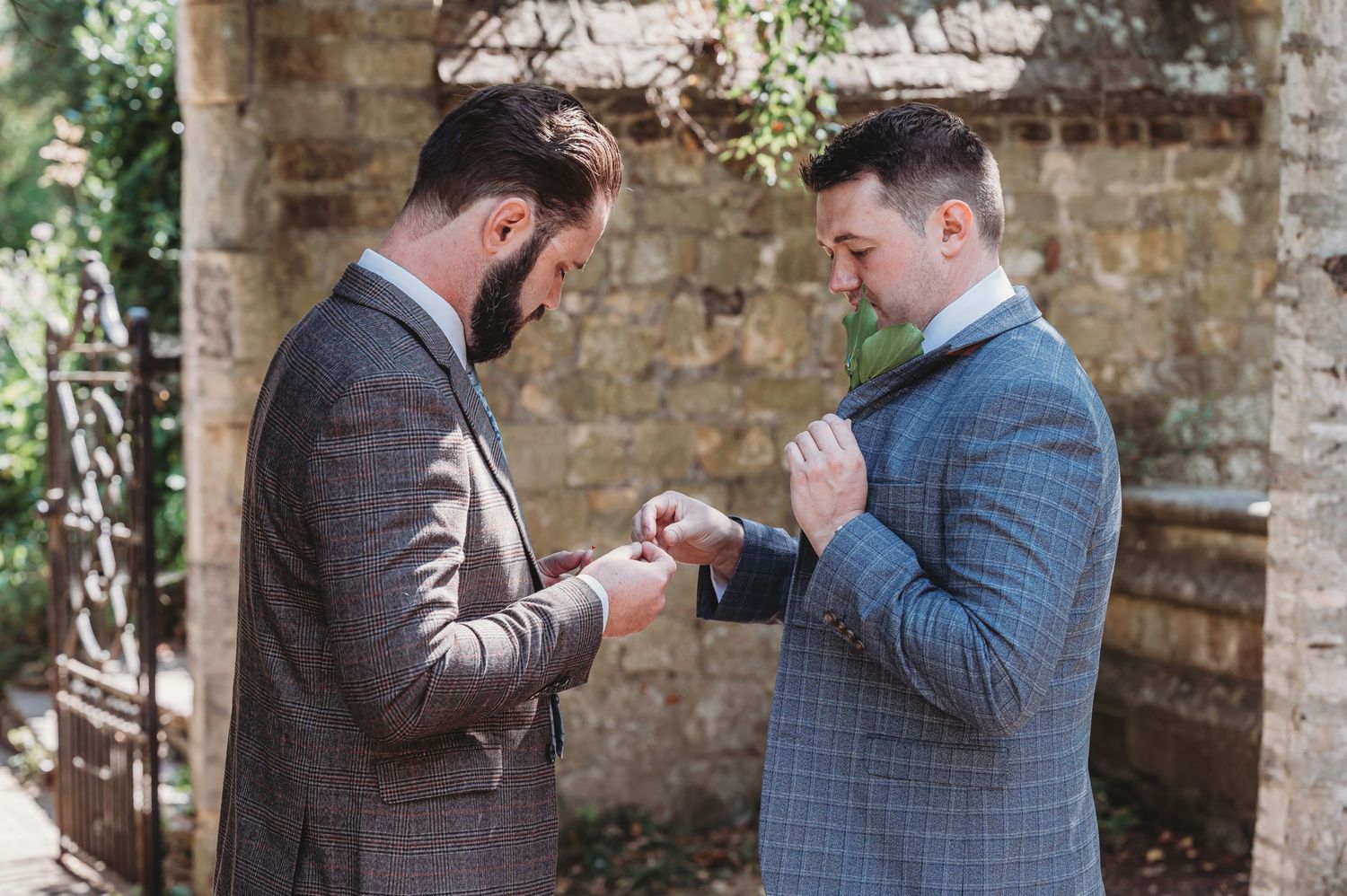 Two people in tailored suits check their watches while standing in front of a rustic stone wall.
