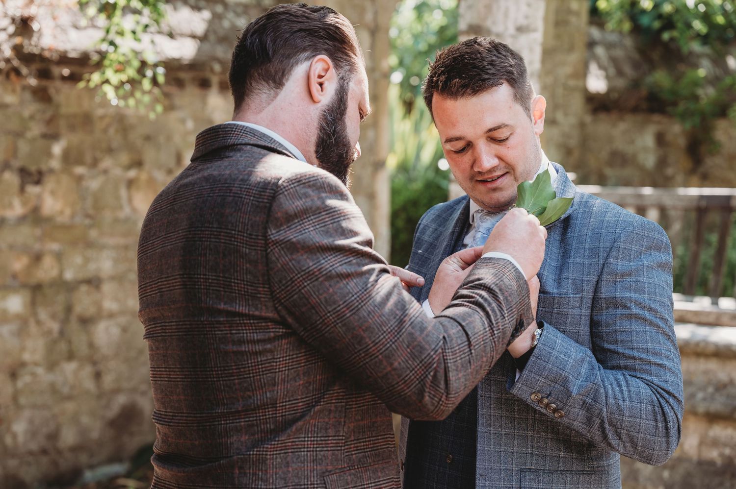 Two well-dressed figures in suits share an intimate moment during a wedding ceremony, adjusting boutonnieres outdoors.