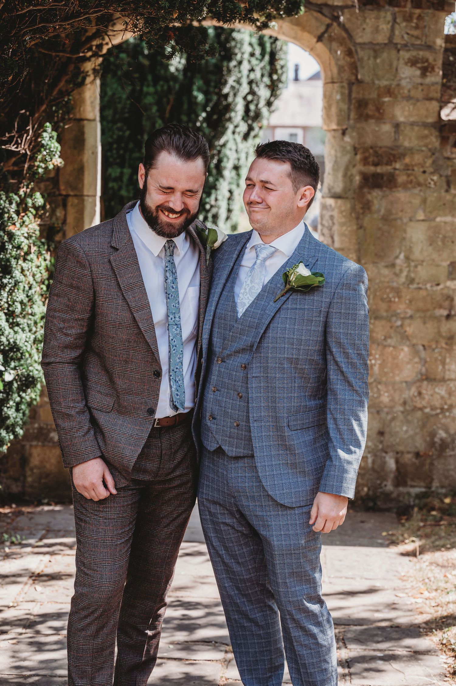 Two people share a joyful moment while wearing stylish suits under a stone archway on a sunny day.