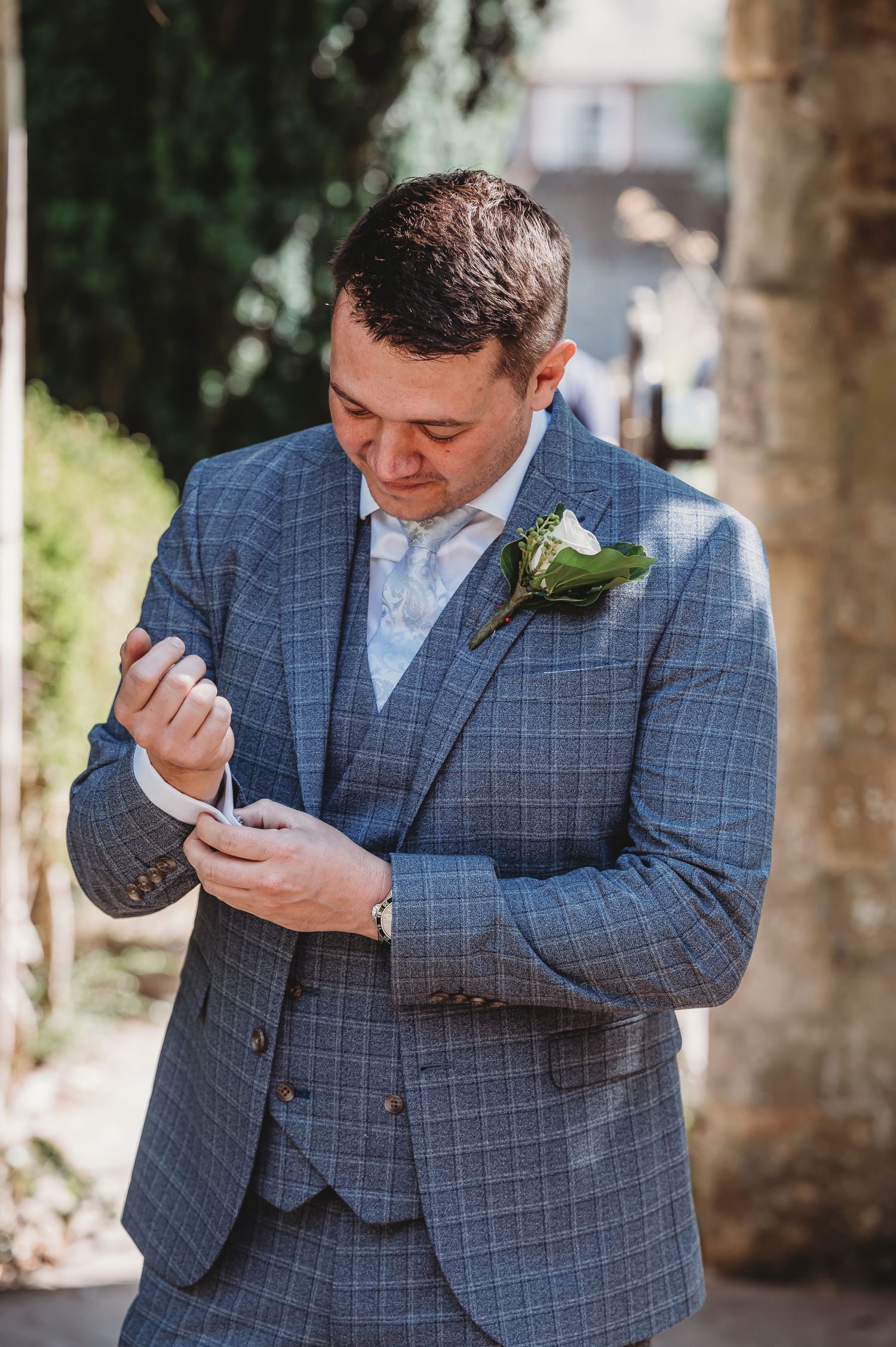 A groom in a blue checkered suit adjusts his cufflinks while wearing a white floral boutonniere at an outdoor wedding.
