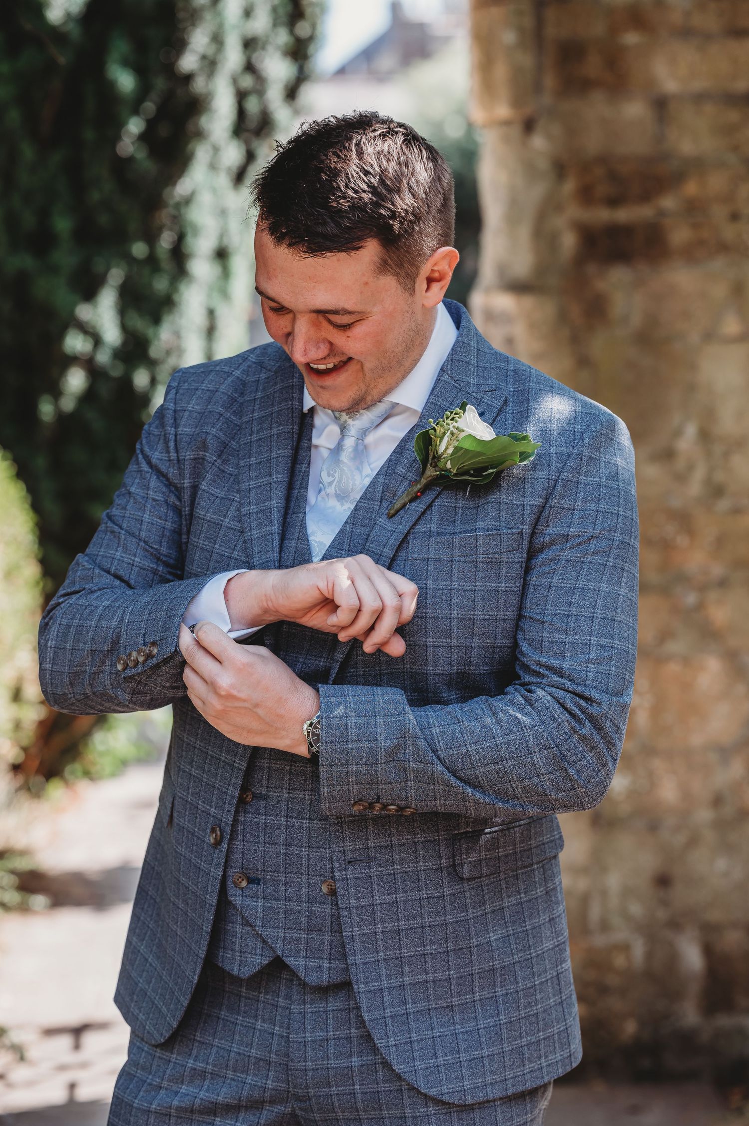 A smiling person in a blue checkered suit and white boutonniere checks their watch during a formal occasion.